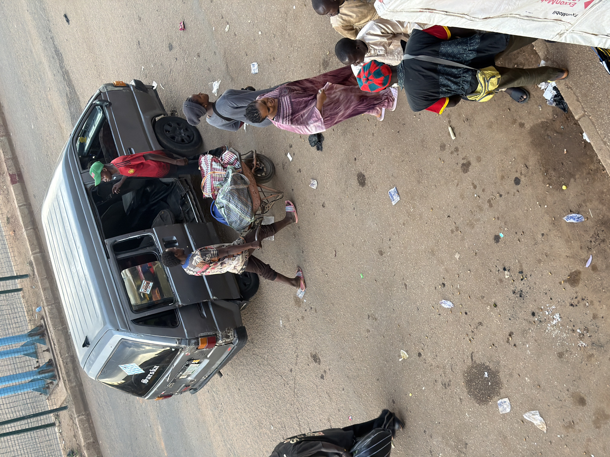The image depicts a street scene in Bukuru, Nigeria, with several people gathered around a gray van. The van appears to be stationary, possibly waiting for passengers or for its next journey.
Several individuals are visible. One person, wearing a red shirt and green cap, is leaning into the open door of the van. Another person, in a purple and white patterned dress, is standing near the van, holding a piece of paper. A person in a gray hoodie and a beanie is also present. Further back, a group of people are seated or crouching, some wearing traditional attire. One person is wearing a black outfit with red accents and a red and green patterned hat. A yellow bag is slung across their shoulder.
In the foreground, a colorful striped bag or mat is placed on the ground, next to what appears to be a handcart. There are also scattered pieces of trash on the ground, including paper and plastic wrappers.
The lighting suggests it could be daytime, possibly late morning or afternoon, with overcast or diffused sunlight. The road surface is asphalt, and there are some blue metal structures and a fence visible in the background, suggesting an urban or semi-urban environment.
Visible text includes "Sarsika" on the back window of the van, and "LAGOS" on a sticker inside the van. There's also some text on a white object in the background that is partially obscured but appears to be a product label, possibly mentioning "ExxonMobil". The overall atmosphere seems to be one of everyday activity, perhaps related to transportation or market vending.