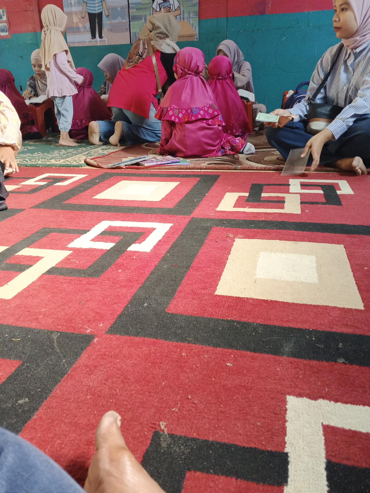 A group of people, mostly young girls, are sitting on the floor in what appears to be a room with a large, red, black, and white patterned carpet. Some of the girls are wearing hijabs and colorful clothing. One young girl in a pink hijab and dress is standing and looking down. In the background, there is a wall with what looks like posters or artwork depicting figures. A woman in a striped shirt and hijab is seated on the floor, holding a clear plastic sheet and some small bills. The scene suggests an indoor gathering, possibly a religious or educational event, in Warungkiara, Indonesia.