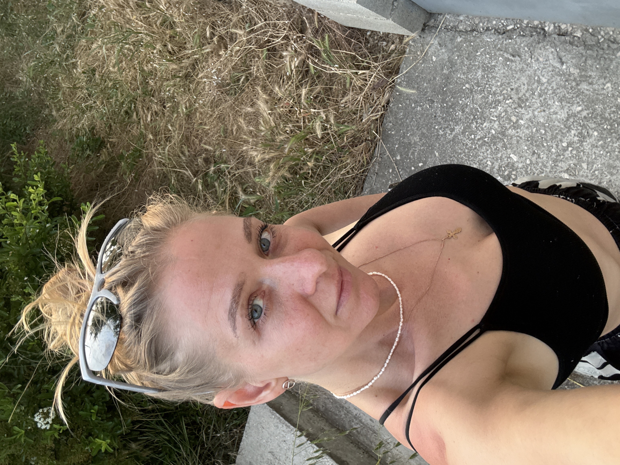 A blonde female is captured in a high-angle selfie in the city of Unknown, Unknown. Her hair is pulled into a messy bun, with loose strands framing her face. Silver-framed, reflective sunglasses rest atop her head. Her blue eyes are directed towards the viewer. A small silver-colored circular earring is present on her visible left ear. She wears a choker of small white pearls and a longer, thin gold-colored chain with a small cross pendant. She wears a black spaghetti-strap crop top, revealing her midriff. A red patch is visible on her left shoulder, consistent with sunburn. The lower edge of black and white patterned clothing is visible at the bottom of the frame. The scene is outdoors, with the subject standing on a light grey, speckled concrete surface. To her left, a light grey concrete ledge or wall is visible, behind which is dense green foliage featuring small white flowers. To her right and in the background, a patch of dry, light brown grass and weeds is present, interspersed with green stalks and small white seed heads.