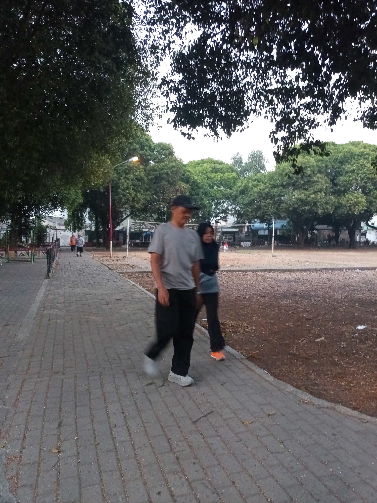 In the city of Bekasi, Indonesia, on a paved walkway with intermittent leafy trees and sparse brown ground cover, two people are walking. A man, wearing a grey t-shirt, dark pants, and a black baseball cap, is walking towards the foreground. Next to him, a woman in a dark top and light-colored pants, also walking towards the foreground, is partially visible. In the background, two more individuals are walking away from the foreground, further down the walkway. A lamppost with a lit bulb stands to the left of the walkway. Beyond the walkway, an open field and some buildings are visible.