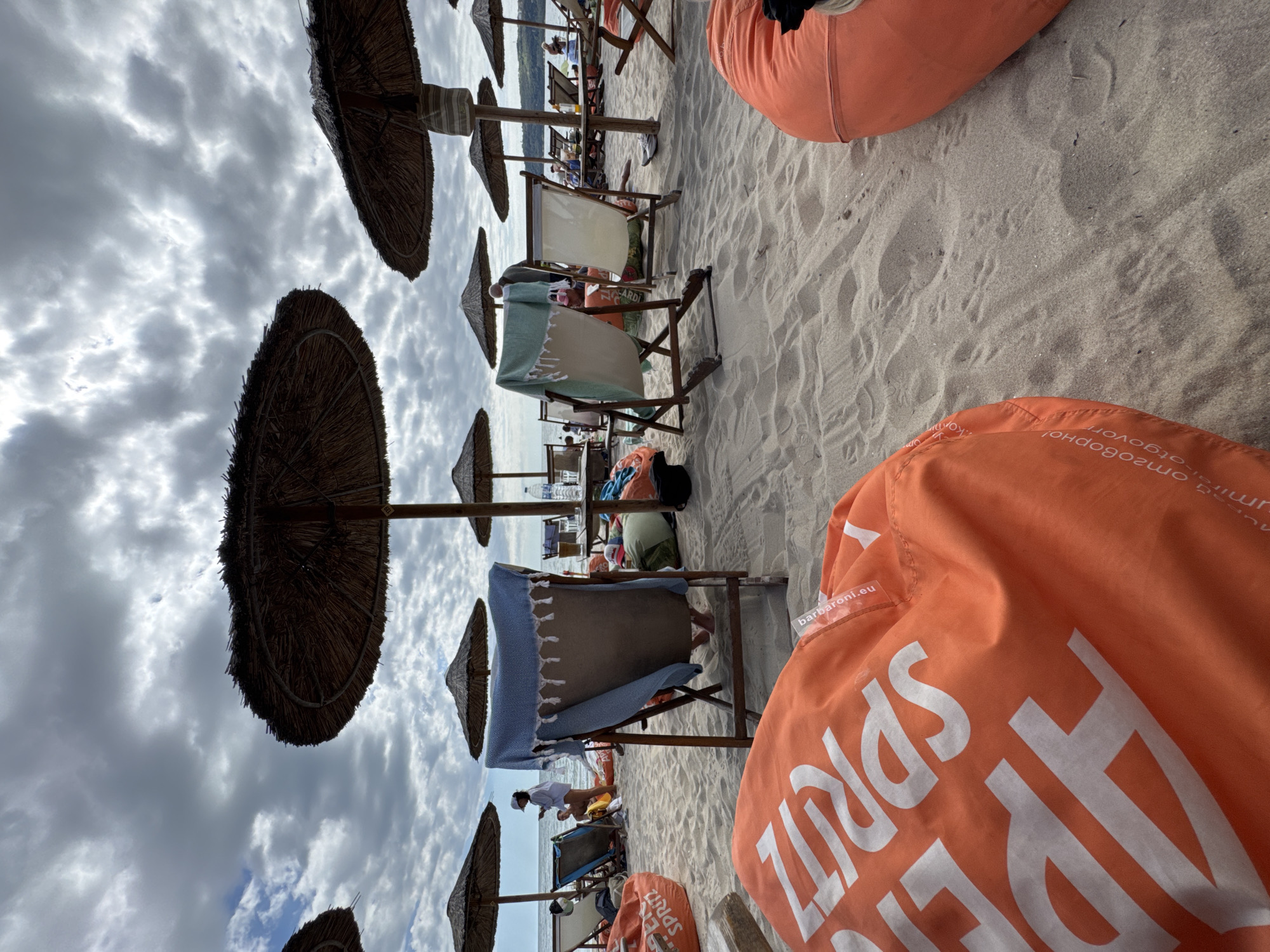 The image depicts a crowded beach scene on a cloudy day at the Golden Sands resort in Varna, Bulgaria. In the foreground, two large, orange beanbag chairs are visible, one of which has white lettering on it. The beach is covered in sand, and several straw umbrellas provide shade for lounge chairs and tables arranged on the sand. In the background, the sea is visible, and several people are scattered across the beach, some sitting on chairs, others standing. The overall impression is of a lively beach resort despite the overcast sky.