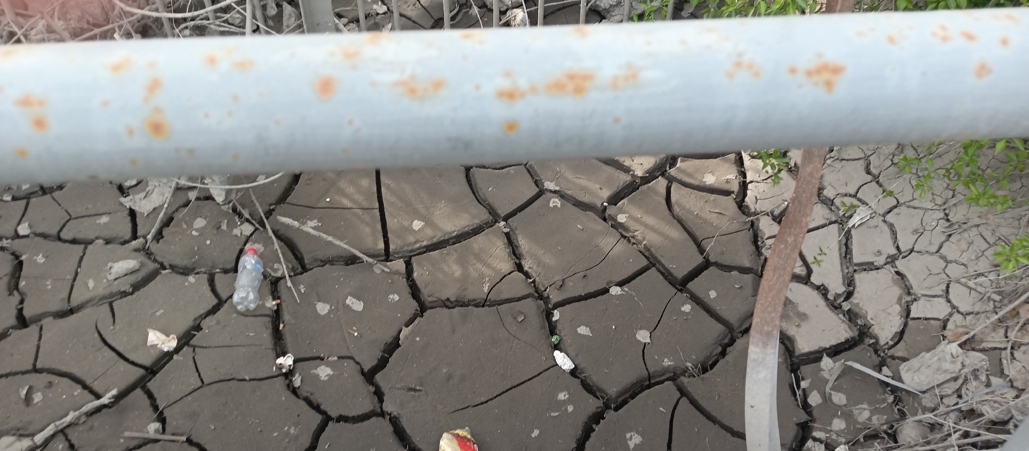 The image depicts a patch of cracked, dry mud, likely at the bottom of a drained body of water. The mud is gray-brown in color, and the cracks create a pattern of irregular polygons. Debris, including a plastic water bottle, a small piece of silver foil, and other miscellaneous trash, is scattered across the cracked surface. In the foreground, a rusted metal pipe or railing obscures the view. Some green foliage is visible on the right side of the image. The location is Unknown.