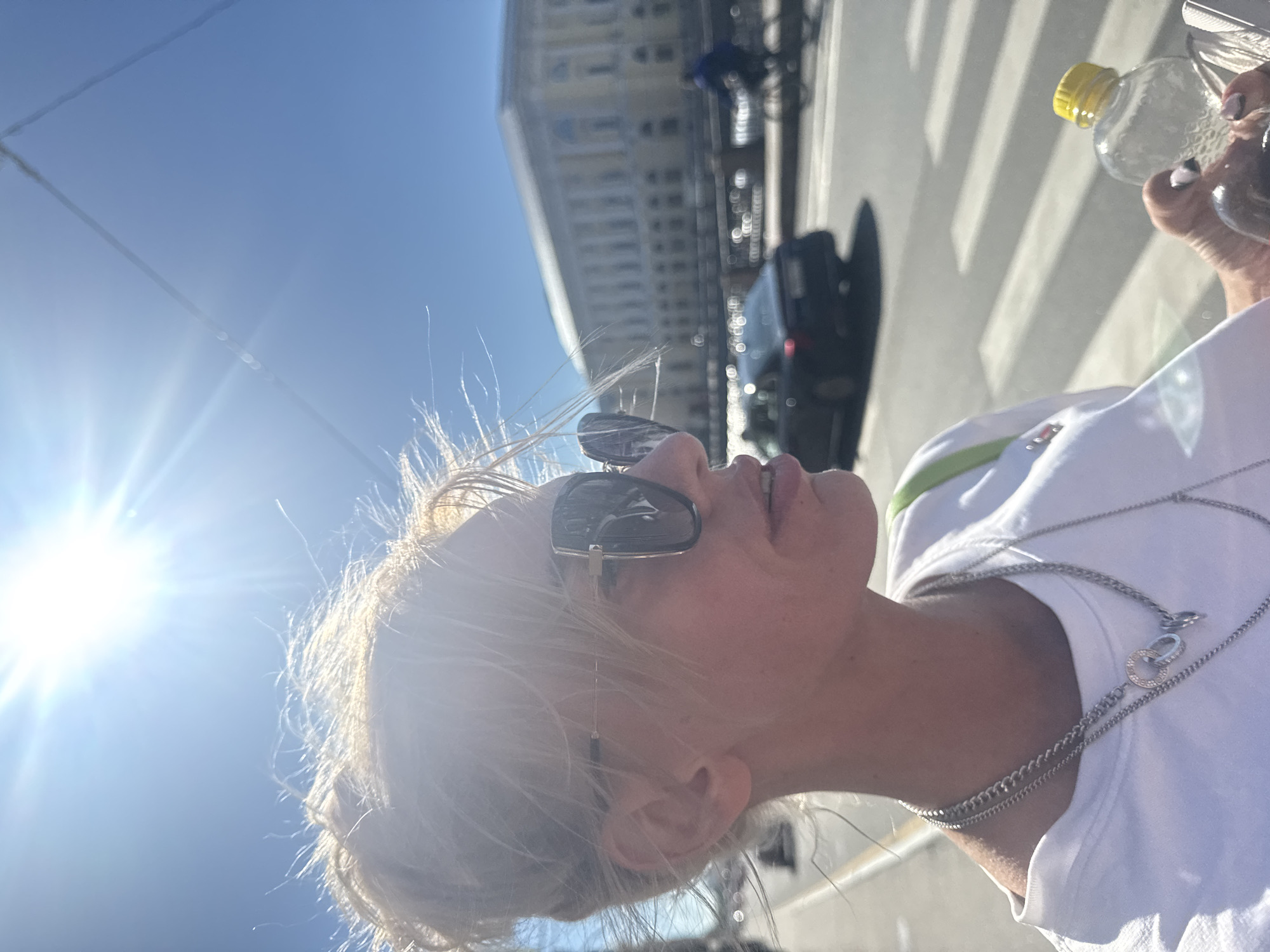 In Saint Petersburg, Russia, a woman with blonde hair pulled back in a bun and wearing sunglasses and a white t-shirt stands outdoors. She is holding a partially full plastic water bottle with a yellow cap in her right hand. Her left hand is not visible. The background features a bright blue sky with the sun shining intensely, creating a lens flare effect. A wide street with a pedestrian crossing is visible, along with a dark-colored car and a person on a bicycle in the distance. A large, light-colored building with columns and multiple windows lines the far side of the street, suggesting an urban setting, likely along a river embankment given the architectural style typical of Saint Petersburg.