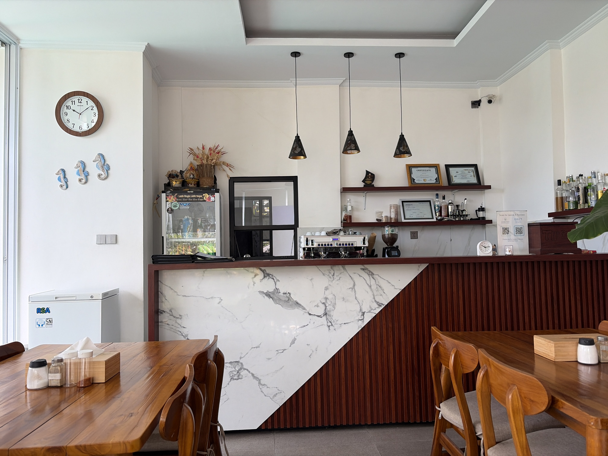 The image depicts the interior of a cafe, likely in Kubu, Indonesia, with a focus on the counter area and a dining table. A wooden dining table with chairs is in the foreground, set with a tissue box and small jars of condiments. Behind the table, a counter with a white marble front and wooden paneling spans across the image. On the counter, a coffee machine, a coffee grinder, and various bottles are visible. Shelves above the counter hold framed certificates and decorative items. A refrigerator is positioned to the left of the counter. The walls are white, and the ceiling features recessed lighting and pendant lights. A clock hangs on the wall to the left, along with decorative seahorse figures. A security camera is mounted on the upper right wall. The overall impression is a clean and modern cafe environment.