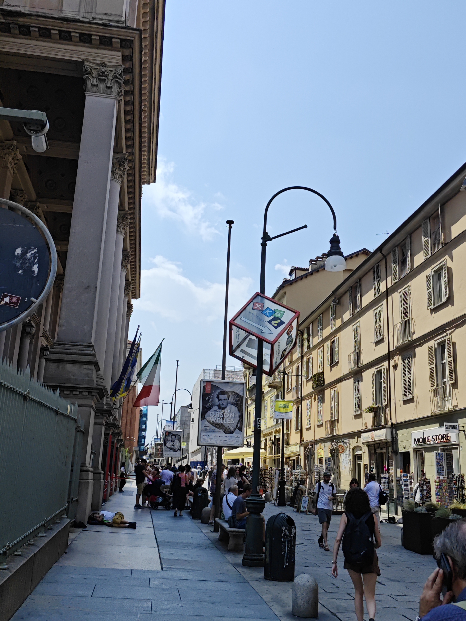 The image depicts a street scene in Turin, Italy, on a sunny day with a clear blue sky. The viewpoint is from a pedestrian walkway bordering a building on the left with classical architecture featuring large columns and an ornate cornice. On the right side of the street, a row of light-colored, multi-story buildings with numerous windows and balconies lines the walkway.
Several people are present, walking along the sidewalk, sitting on benches, and standing in groups. One person is lying down on the sidewalk near the left building. A street lamp with a curved arm and a light fixture extends over the walkway. A translucent, cube-shaped sign with colorful symbols and text is suspended from the lamp post.
Various flags, including the Italian flag, are displayed along the left building. Posters are visible on the right side of the street, one of which appears to be a movie poster featuring Orson Welles. Shops with awnings and displays of merchandise are situated on the ground floor of the buildings on the right. The walkway is paved with stone tiles. The overall impression is of a busy urban street, likely a commercial area.