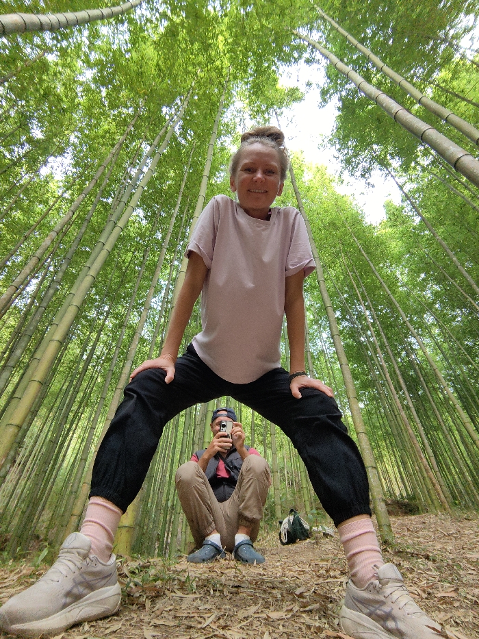 The scene is set within a dense bamboo forest in Mù Cang Chải, Vietnam. A light-skinned adult female stands prominently in the foreground, facing the camera with a slight smile. She wears a light pink short-sleeved t-shirt, black loose-fitting pants, pink socks, and light-colored athletic shoes. Her legs are spread wide and knees bent, with her hands resting on her thighs. Positioned directly beneath her, seated in a low crouch between her legs, is a light-skinned adult male. He wears a dark baseball cap backward, a dark vest over a reddish t-shirt, light brown pants, and blue slip-on shoes. He holds a white smartphone horizontally with both hands, aiming it upwards, appearing to take a photograph. A dark green bag with white netting rests on the ground to his right. The environment consists of numerous tall, slender green bamboo stalks extending upwards, creating a dense, verdant canopy against a bright sky. The forest floor is covered with dry leaves and natural debris. The image is captured with a wide-angle, low perspective.