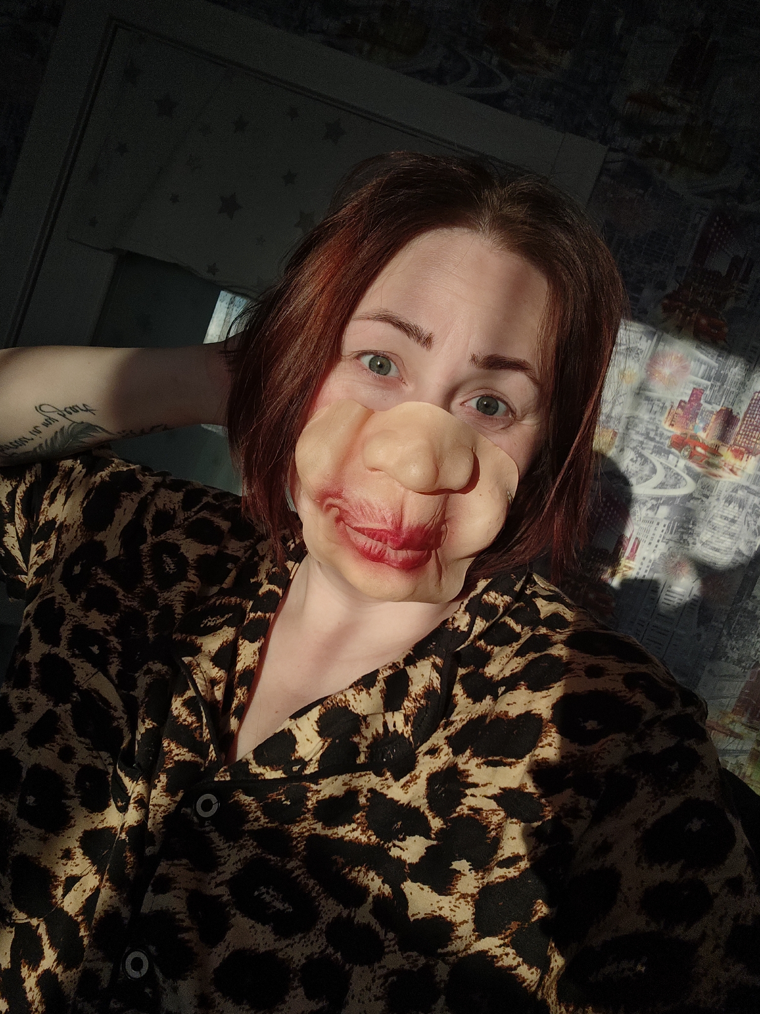 A woman with fair skin and auburn hair is seen in an indoor selfie. She is wearing a leopard-print shirt and holding her hand behind her head. She is also wearing a novelty prosthetic mask resembling a large nose and lips painted with red lipstick. The background includes a dark-colored wall on the left and a patterned wall or bedspread on the right. The image is partially illuminated by sunlight, creating shadows and highlights throughout the scene. This photo was taken in Markovskii, Russia.