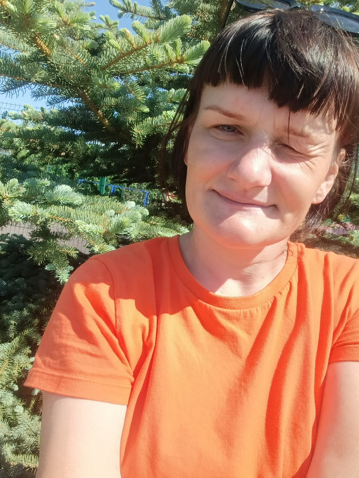A woman with short, dark brown hair and an orange t-shirt is winking at the camera. She is standing outside, with a large evergreen tree in the background. The sky is visible through the tree branches. The picture appears to be taken in bright sunlight. There is no indication in the image that it is Фоки, Russia.