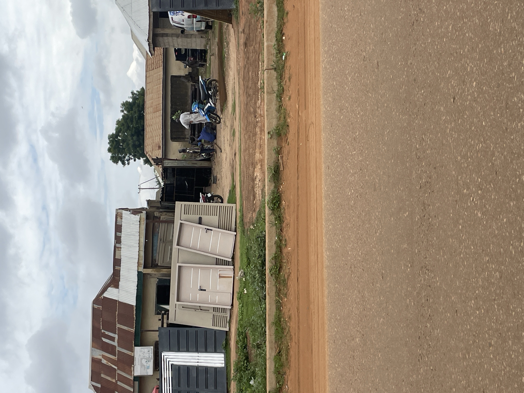 The image captures a scene in Minna, Nigeria, likely a workshop or storefront displaying metal doors and gates. Several large, light-colored metal doors are propped up in the foreground, leaning against each other and a wall. To the left, a stack of dark, modern-looking gates or panels is visible. In the background, several people are gathered around a blue motorcycle, suggesting activity related to either repairs or sales. One person, dressed in white, stands near the motorcycle, while others are seated or standing nearby. Behind them, a modest building with a corrugated metal roof is visible, alongside other structures and a white van. The environment appears to be an open area, possibly a street or yard, with dirt and a concrete barrier in the foreground. The sky is overcast.