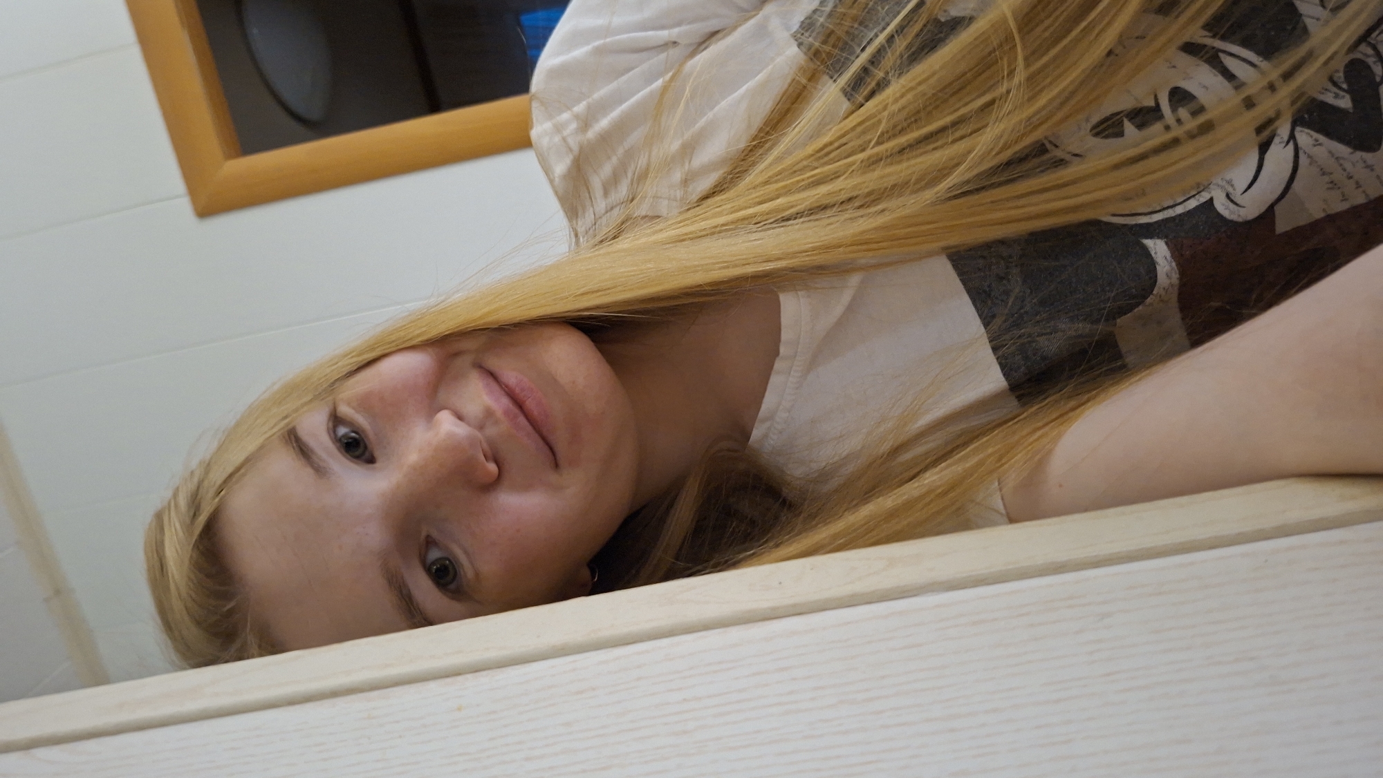 A young woman with long blonde hair is leaning against a light-colored wooden doorframe. She is wearing a white t-shirt with a graphic print. Her head is tilted and she is looking at the camera with a slight smile. The background appears to be an indoor setting with a white, paneled wall and a doorway with a wooden frame. The image is a close-up portrait. The location of Большой Букор, Russia is not discernible from the provided image.
