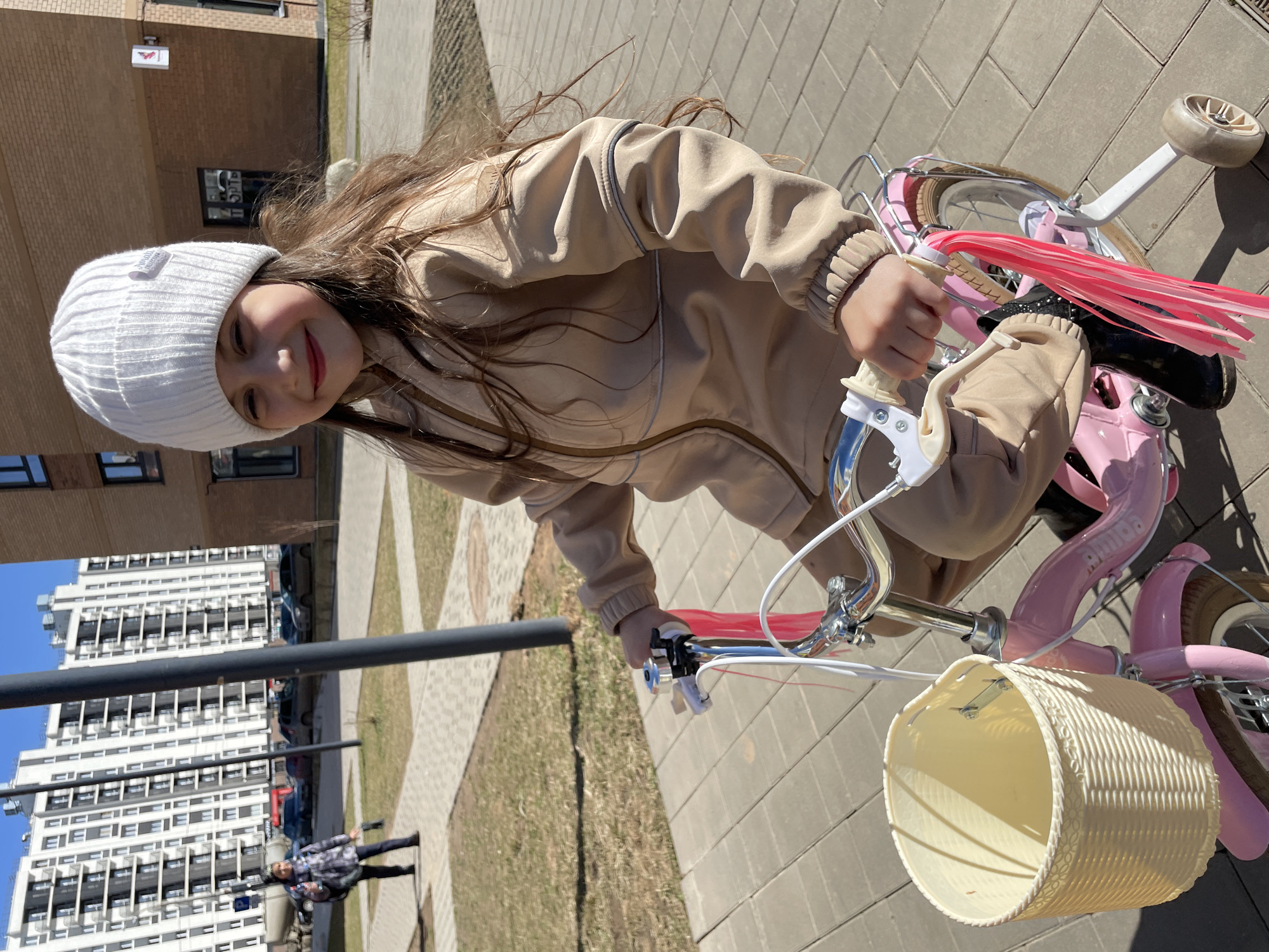 A young girl in a white knitted hat and beige jacket smiles while riding a pink bicycle with a white basket and pink tassels on the handlebars. The scene is outdoors, with a paved walkway and patches of grass visible. A tall apartment building is in the background, with other people walking along the path. The location is likely Izhevsk, Russia.