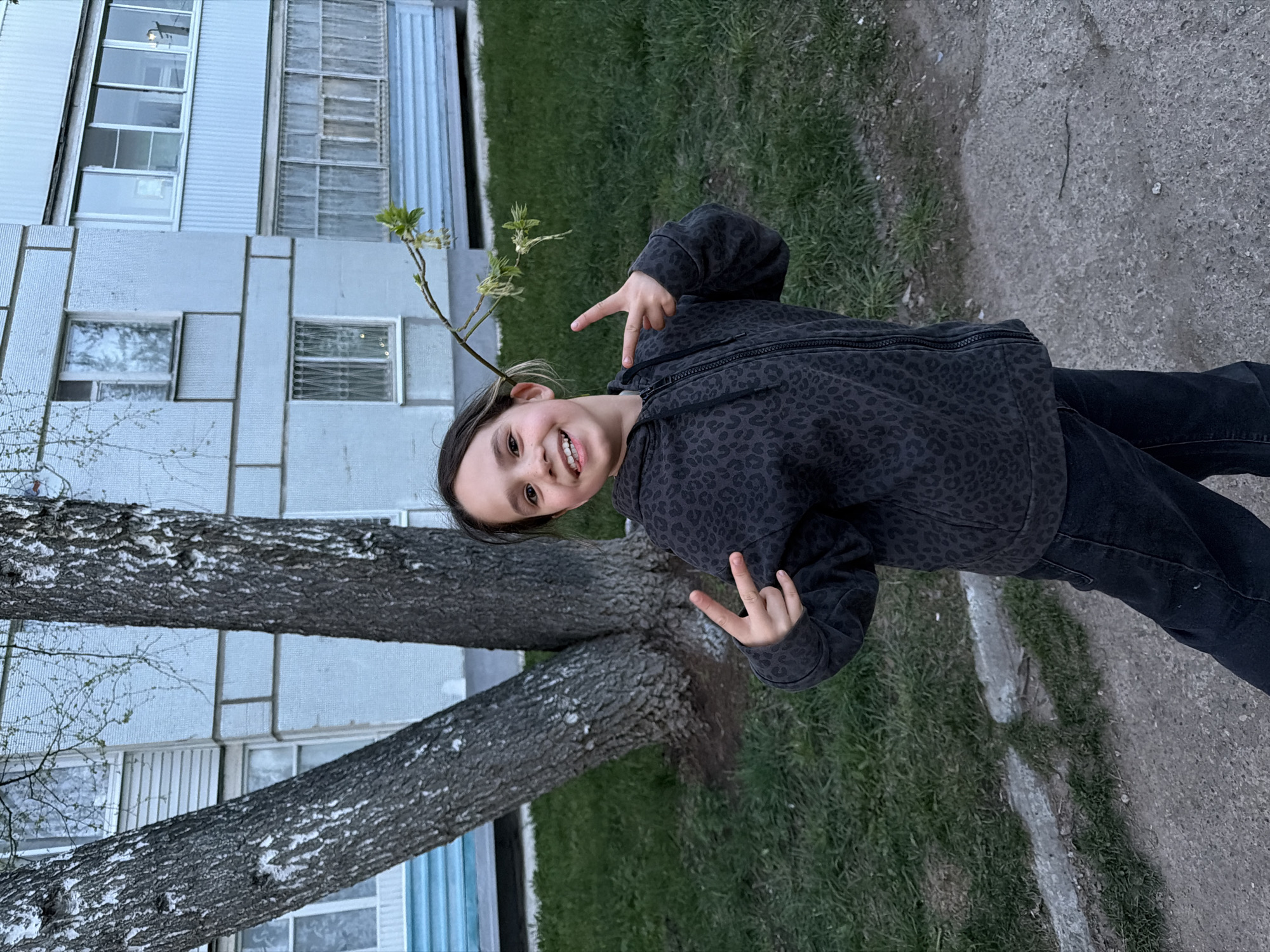 A young girl stands in Chaikovsky, Russia. She wears a dark jacket and black pants. She poses with both hands showing a peace sign. She stands next to two tree trunks. Behind her, is a gray apartment building. The ground shows grass and a paved area.