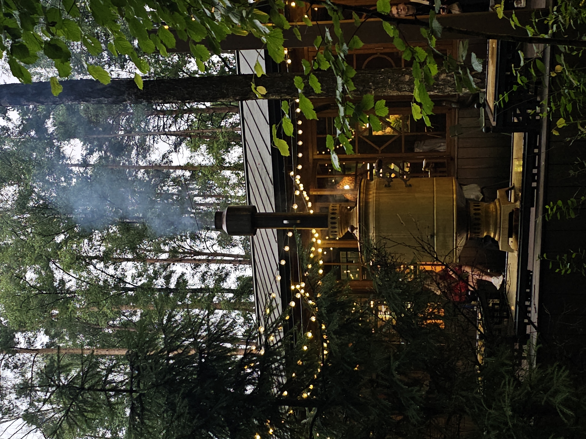 The image shows an outdoor seating area in a forest, likely part of a restaurant or cafe, located in Nemchinovo, Russia. In the foreground, a large, golden samovar is actively emitting smoke, suggesting it is in use. String lights are draped across the scene, illuminating a wooden structure with large windows in the background. One person is visible through the windows, sitting at a table. Another person, a young woman with glasses, stands near the entrance of the structure, looking to the left. The area is surrounded by tall trees, with some of the leaves obscuring parts of the scene. The overall atmosphere is rustic and cozy, with a hint of a chilly evening suggested by the smoke and lighting.