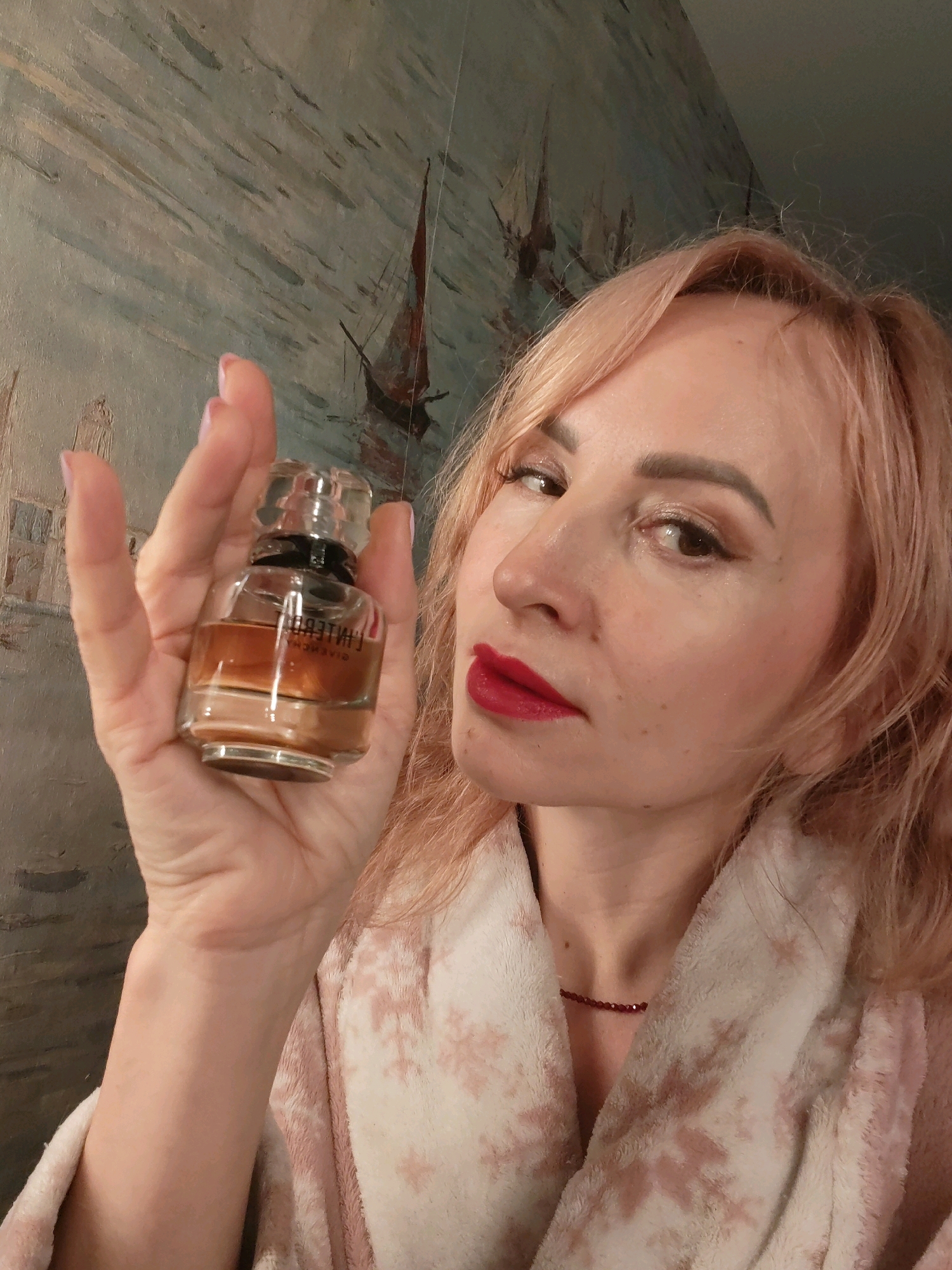 In an indoor setting, a woman with light blonde hair and red lipstick is holding a small glass perfume bottle with a clear, faceted cap. The bottle contains amber-colored liquid. She is wearing a pink bathrobe with a snowflake pattern. Behind her, a painting of ships on water is visible. The lighting suggests an interior space, potentially a home. There is no information about the location in Chaikovsky, Russia.