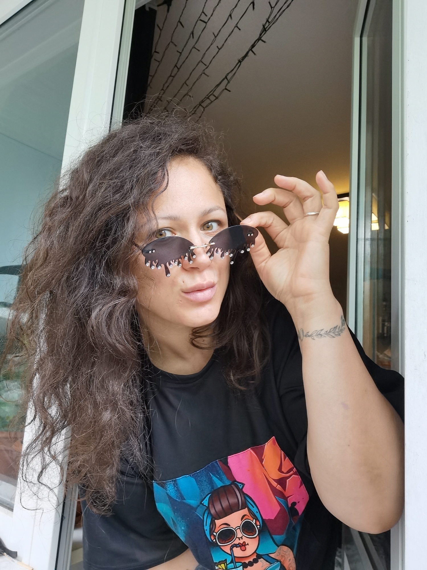 A medium close-up portrait captures a woman with voluminous, dark brown curly hair and light brown eyes, looking directly at the viewer with a slight, confident smirk. She is positioned within a white door or window frame, suggesting an indoor or transitional space in Chaikovsky, Russia. Her right hand is raised, holding up a pair of distinctive dark sunglasses that feature a 'dripping' or 'melting' design, embellished with small clear gems. A simple silver-toned ring adorns her right ring finger.
She is wearing a black t-shirt with a vibrant graphic print on the front. The visible part of the design depicts a stylized cartoon woman with dark hair styled in a bun, large round sunglasses, a pearl necklace, and holding a drink with a straw. Her left wrist bears a delicate tattoo of a leafy or floral wreath. In the upper background, string lights are visible, adding to the ambient setting. The bright lighting suggests it is daytime. No discernible text is present in the image.