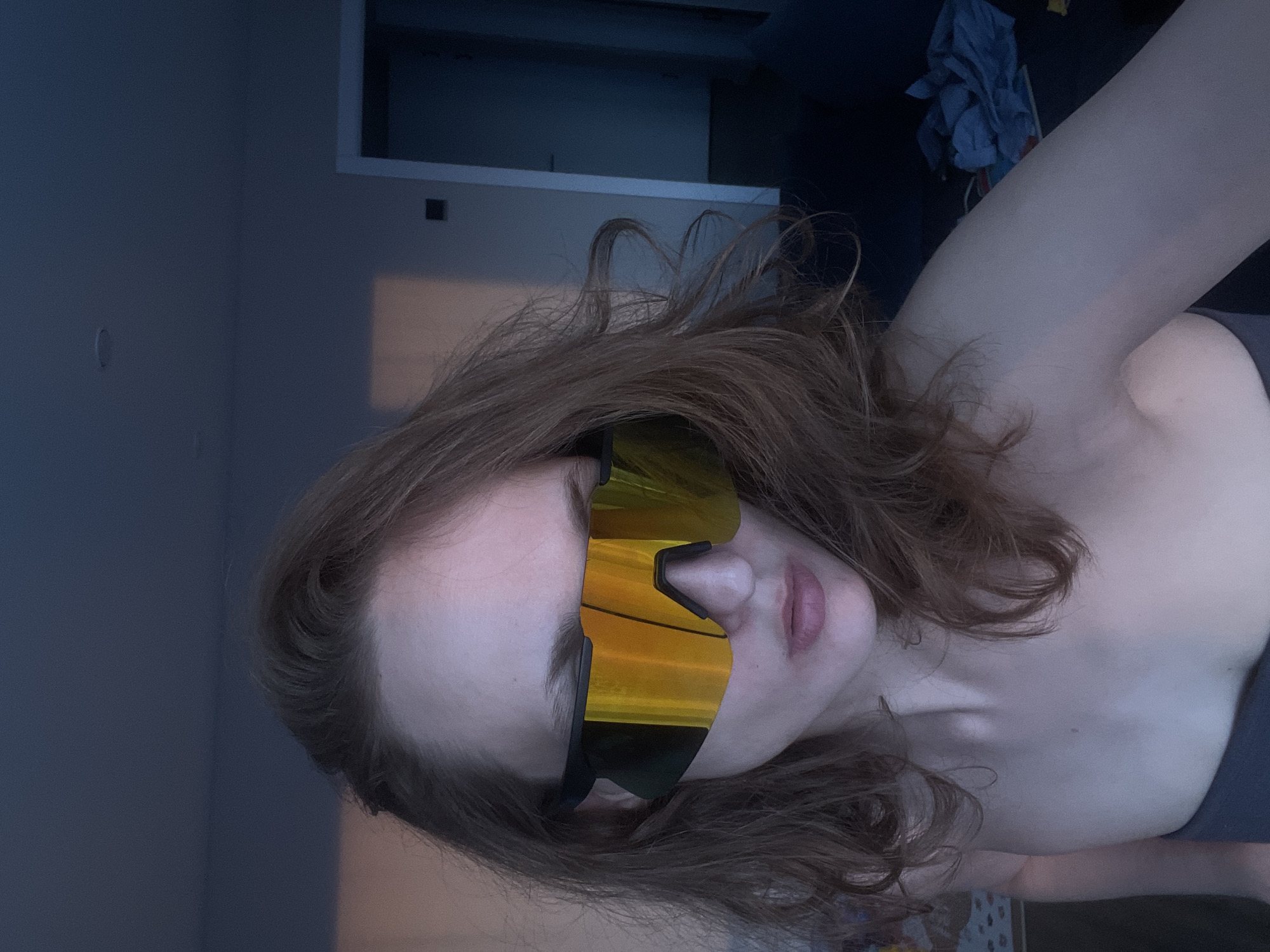 The image features a person's upper body and head, tilted to the side, in an indoor setting in Podshivalovo, Russia. The subject appears to be female, with light skin and long, wavy reddish-brown hair. She is wearing large, wrap-around sunglasses with highly reflective, golden-orange lenses and black frames, which obscure her eyes. Her lips are slightly parted. A dark-colored top or garment is visible on her shoulder and upper chest.
The background is dimly lit, suggesting evening or low light conditions. To the left and upper left, a plain, dark blue-grey wall is visible. Towards the upper right, there appears to be a lighter-colored architectural feature, possibly a doorframe or part of a built-in shelf, against a darker background. Further to the right, some crumpled, dark blue fabric or clothing can be seen on what might be a bed or couch. The overall atmosphere is muted due to the low lighting. There is no visible text in the image. The person is likely posing for a casual photo.