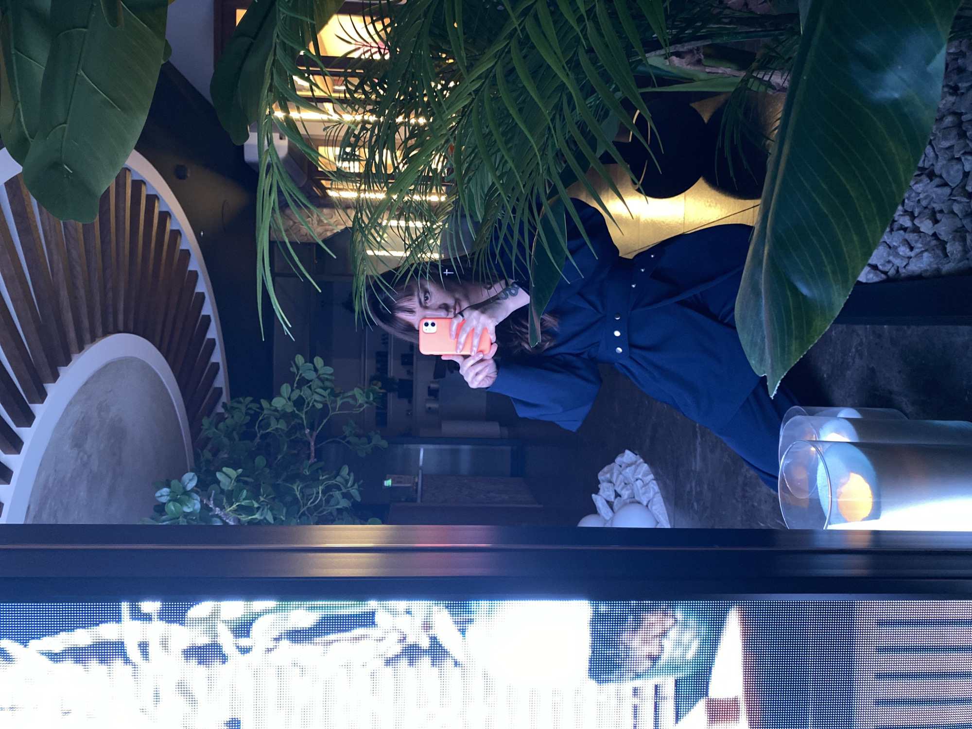 A woman is taking a selfie in front of a mirrored surface. She is holding an orange smartphone. The mirror reflects a partially obscured interior scene with plants, a decorative wall design, and possibly decorative objects. The woman is wearing a dark blue garment with silver button details. An LED display screen is visible below the mirrored surface. The location, Votkinsk, Russia, cannot be verified from the image content.