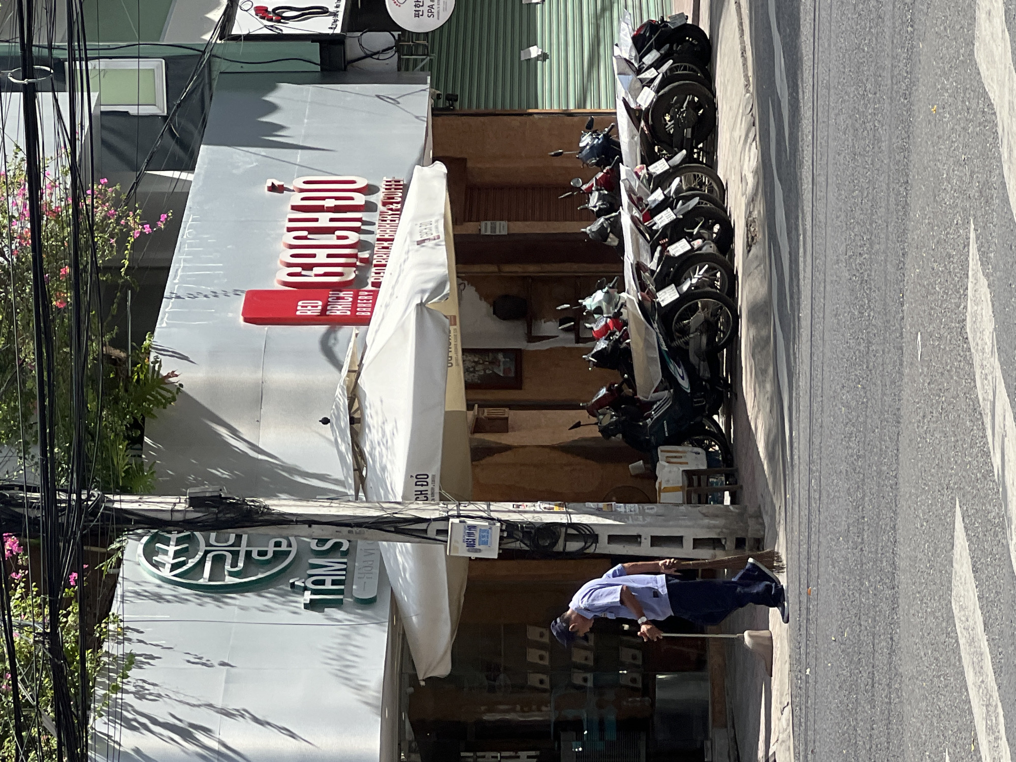 The image shows a street scene in Nha Trang, Vietnam. In the foreground, a man wearing a blue shirt, dark pants, and a cap is sweeping the sidewalk with a broom. He is walking from left to right. Behind him, a row of motorcycles is parked along the curb. A white awning extends from a building that features signage for "RED BRICK BAKERY & COFFEE". The building also has a green logo with Vietnamese text. The street is paved with asphalt and has a pedestrian crossing. Overhead, a tangle of electrical wires is visible against a bright sky. The overall impression is of a typical urban street in Vietnam on a sunny day.