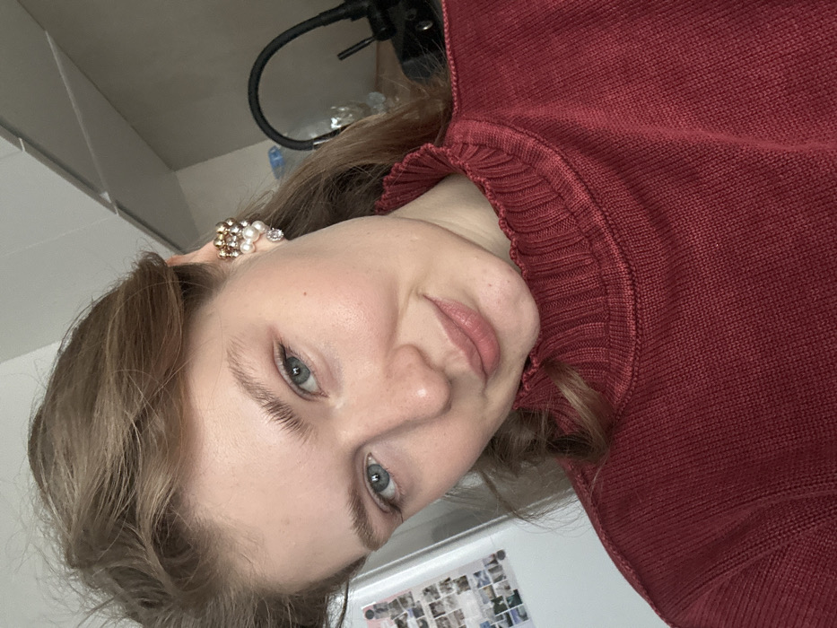 A young adult female is depicted in a close-up, chest-up shot, gazing directly at the camera with a subtle smile. She has light-toned skin, blue eyes, and light brown hair. She wears a deep reddish-brown knit sweater with a distinct ruffled high collar. On her visible right ear, a multi-component ear cuff is worn, featuring a cluster of small golden beads, white pearls, and a clear faceted stone at the bottom. The background on the left reveals a portion of a light-colored appliance, likely a refrigerator, adorned with several small square photographs. To the right, a black gooseneck fixture, potentially a faucet or lamp, and several transparent plastic bottles are partially visible. The scene is situated in an indoor environment within the city of Kazan, Russia.
