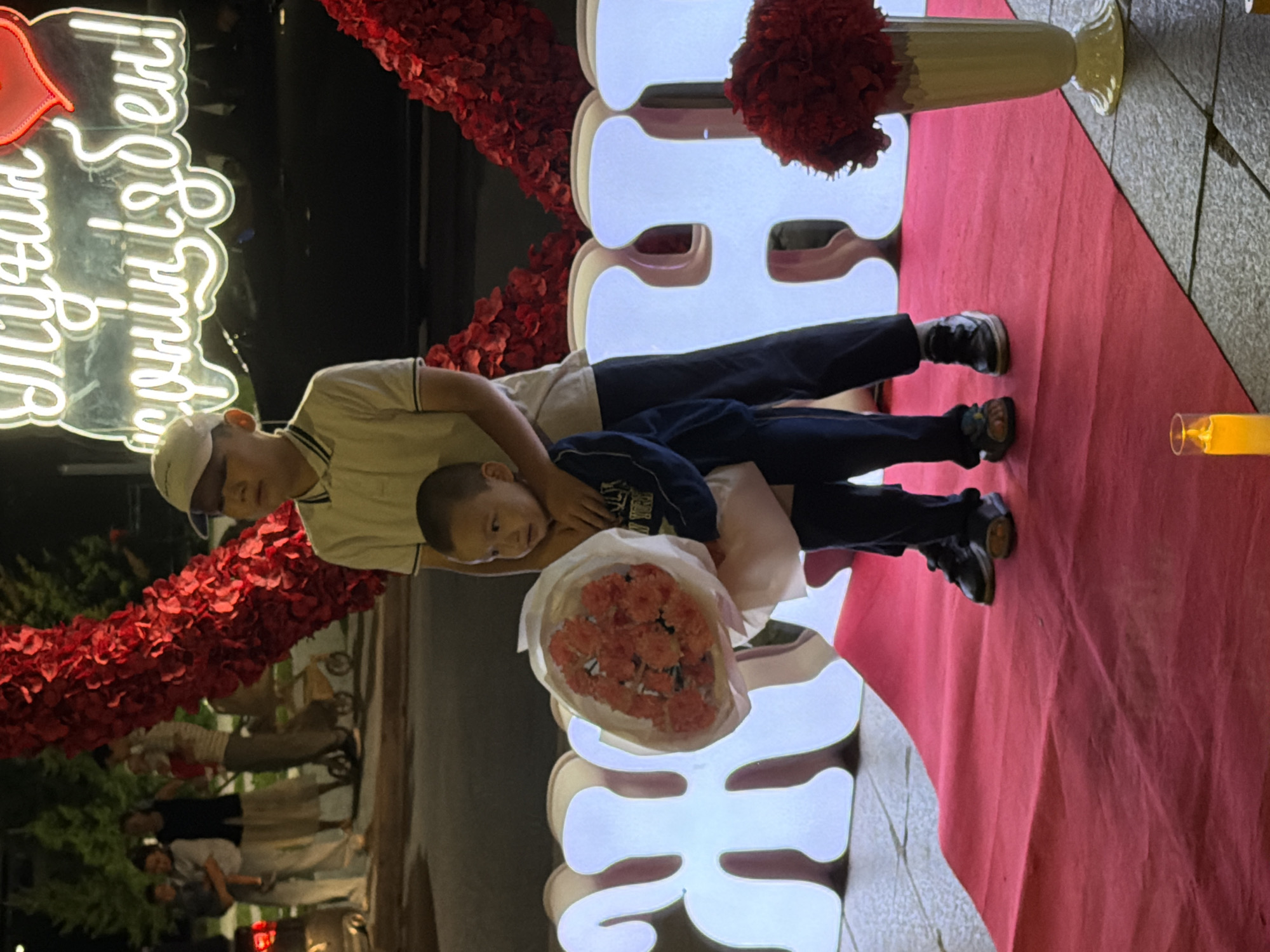 The image captures a scene on a red carpet in Astana, Kazakhstan. Two young boys are the central focus. The older boy, wearing a white cap and a collared shirt, stands behind and slightly to the side of the younger boy. The younger boy, dressed in a dark long-sleeved shirt and dark pants, holds a bouquet of pink flowers wrapped in white paper. Behind them, large, illuminated white letters spell out a word, with a neon sign with writing and a heart above them. The background shows a decorative archway adorned with red petals and foliage, typical of event decorations. Further in the background, several adults and a baby are visible, along with a stroller and a car. The setting appears to be an outdoor public space in Astana, possibly during an evening event.