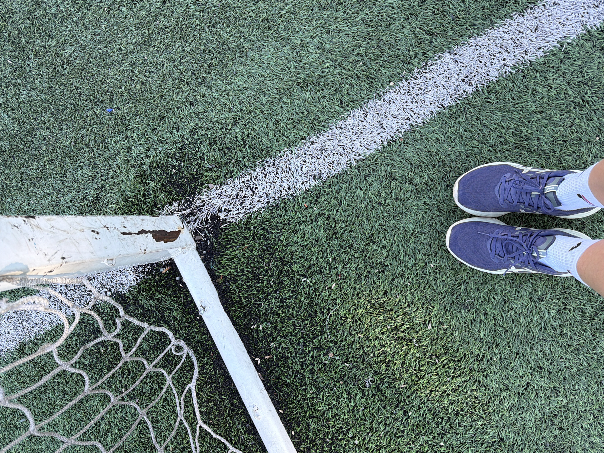A ground-level photograph depicts a section of an artificial turf soccer field in Gorno-Altaysk, Russia. A portion of a white-painted goalpost and net is visible in the lower left corner. A white line crosses the upper left quadrant of the frame. Two feet, shod in navy blue athletic shoes and white socks, are positioned in the upper right quadrant of the frame.