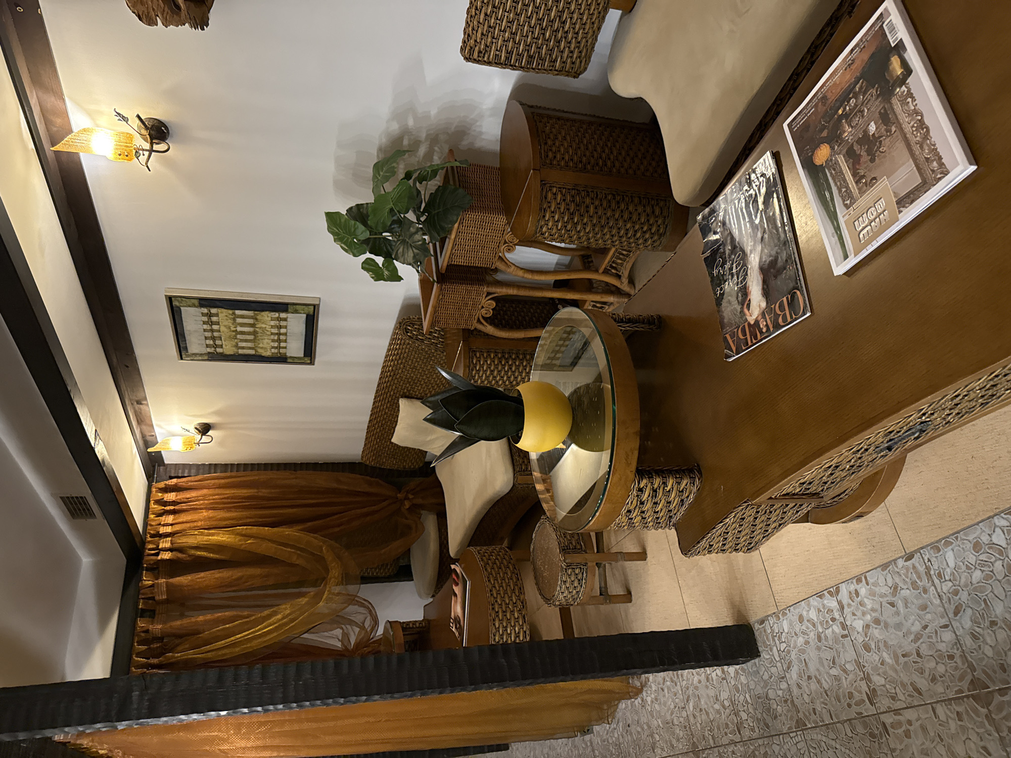 The image depicts the interior of a room, possibly a waiting area or lounge, with a distinct "spa" or "oriental" aesthetic. The room features furniture made of woven rattan and wood, including a sofa, chairs, and tables. The color palette is warm, dominated by shades of brown, orange, and beige, with pops of green from a potted plant.
On the wall, a framed artwork with geometric patterns and a unique sconce light fixture are visible. Sheer orange curtains hang across a window or opening, adding to the ambiance. Two magazines are placed on a wooden coffee table.
The flooring is a mosaic tile pattern in beige and grey tones. The overall impression is one of a tranquil and inviting space. The scene is static, with no people or animals present. The image is captured from a slightly elevated angle, looking down onto the furniture and floor.
While the prompt requests information about the location in Bansko, Bulgaria, there are no visual cues within the image to confirm this specific location. The description provided is solely based on the visual elements present in the photograph.