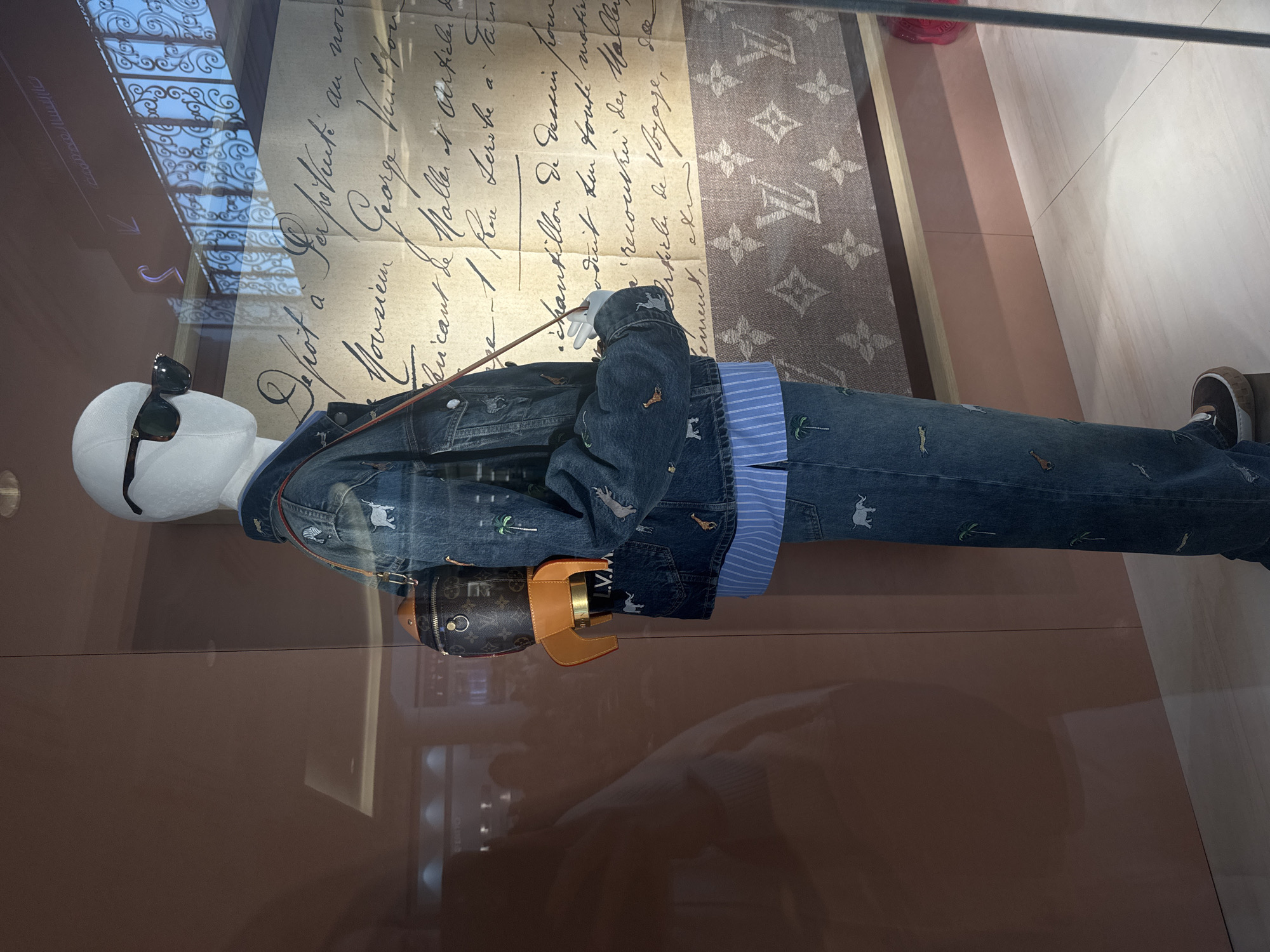 The image is a close-up shot of a mannequin dressed in Louis Vuitton clothing inside a retail display, potentially in Dubai, United Arab Emirates. The mannequin is wearing a denim jacket with animal embroidery over a blue striped shirt and matching denim jeans with animal embroidery. A small, rocket-shaped Louis Vuitton bag is slung across the mannequin's body.
The mannequin has a white head with sunglasses. The background includes a large paper with old-style handwriting and a Louis Vuitton monogram pattern. The retail display is in a well-lit area with a light-colored floor. The display is within an environment reflecting the modern design typically found in Dubai.