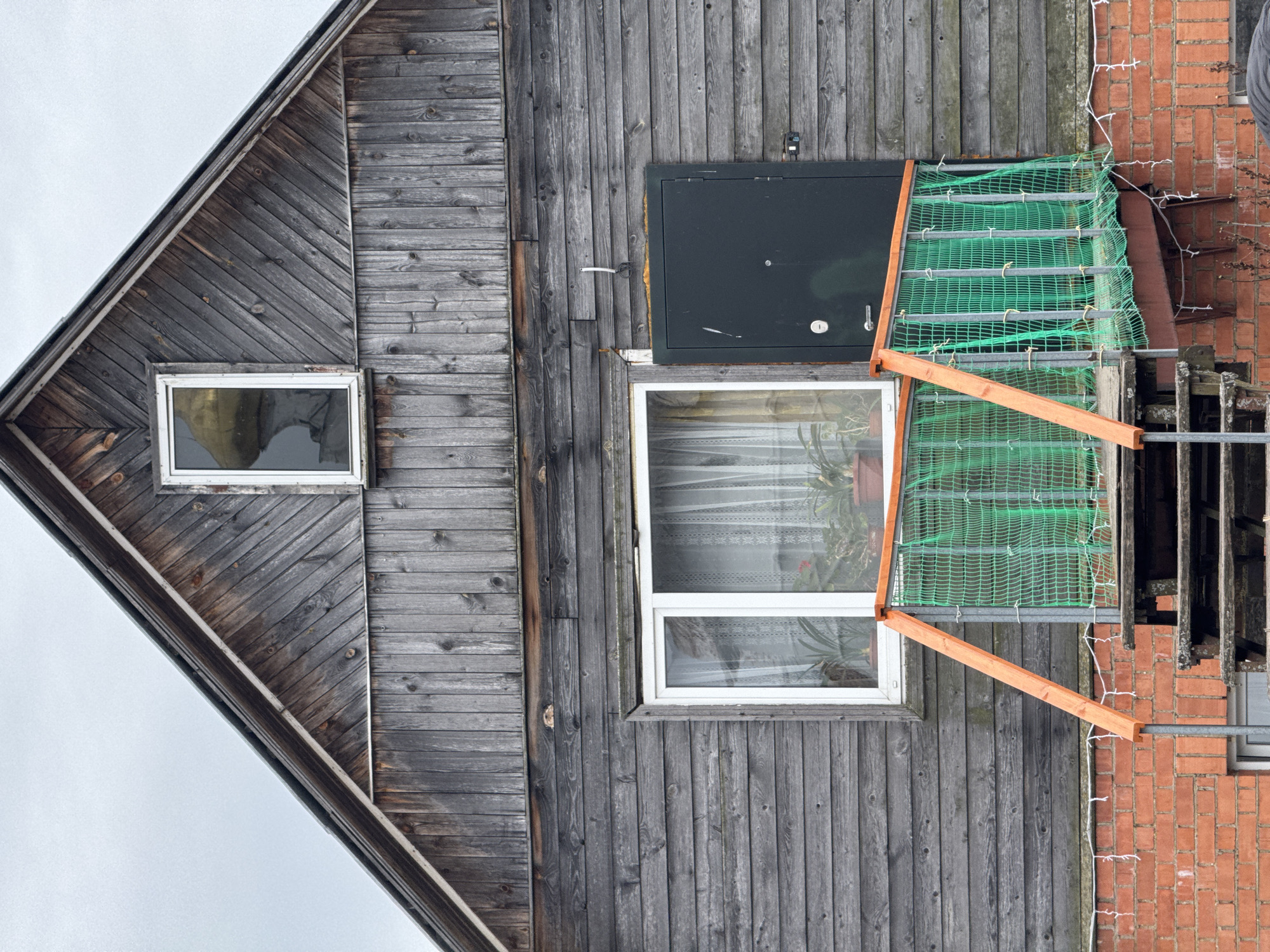 A photograph shows a gray, two-story wooden building with a gabled roof. The left side of the building has diagonally-laid wood siding, with a single small window. The right side has vertical wood siding. Above the right window is a black object. A makeshift balcony or platform covered with green netting extends from the right window, appearing structurally unstable. A brick building is visible to the right of the wooden structure. The image is taken from a low angle, with gray sky visible. There is no indication of the city of Ketoshi, Russia. There are no people in the image.