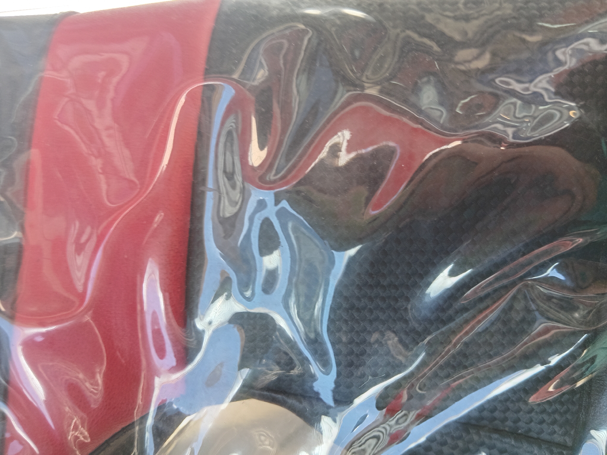 The image shows a close-up of an item, likely a car seat cover, still in its plastic packaging. The packaging creates distorted reflections of the surrounding environment. The cover appears to have a red section and a black section with a textured or patterned surface. The reflections in the plastic suggest an outdoor scene, possibly with buildings and the sky visible. The exact location in Batumi, Georgia cannot be determined from this image alone, as the reflections are too indistinct to identify any specific landmarks.