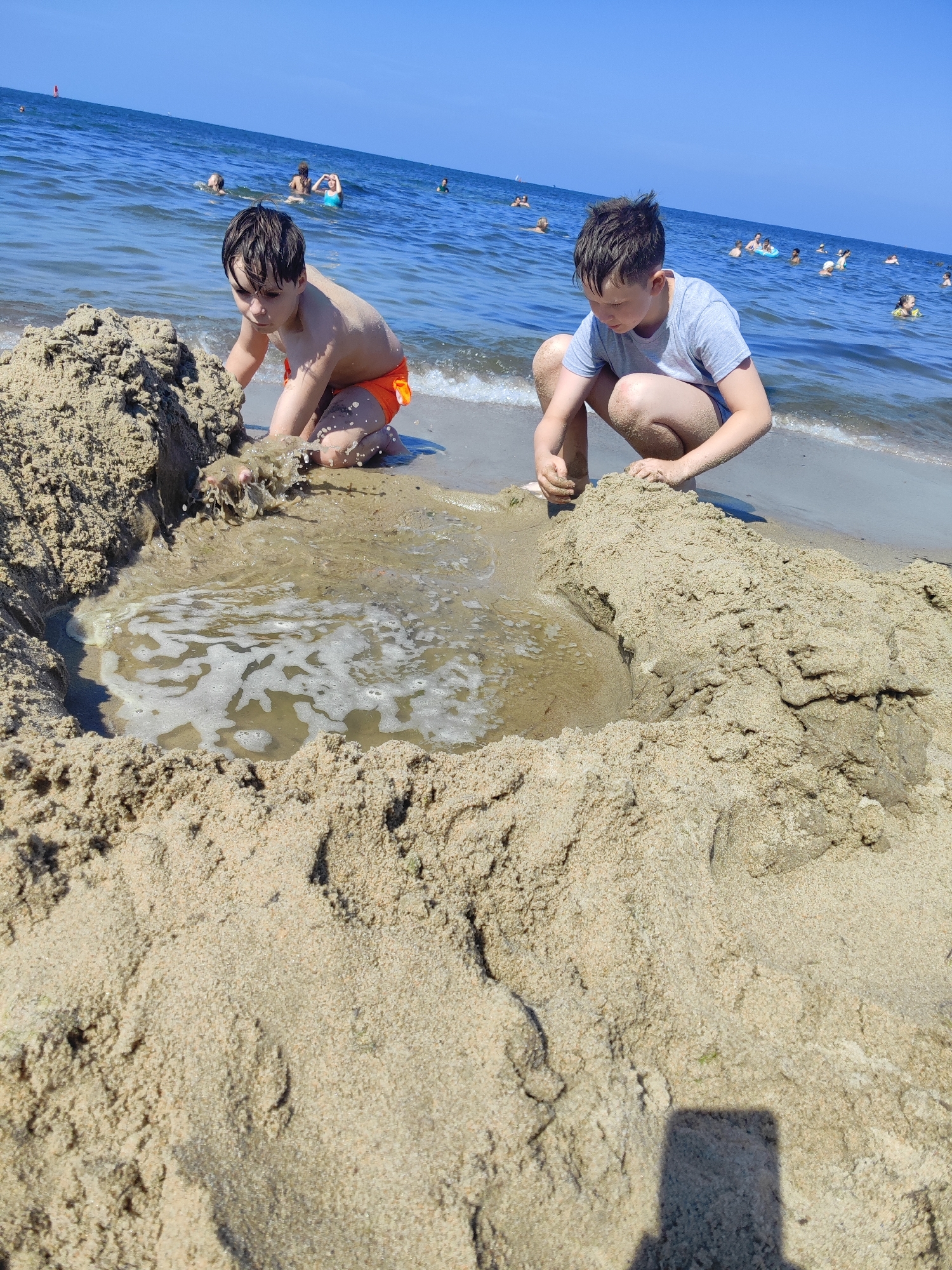 In Pionersky, Russia, on a sunny day at the beach, two young boys are building a sandcastle near the ocean. The boy on the left, wearing orange swim trunks, is kneeling and digging in the wet sand, causing a splash. The boy on the right, wearing a grey t-shirt and blue swim trunks, is also crouching and playing in the sand. A shallow pool of water has formed in front of them, created by the tide. The ocean stretches out behind them, with several other people swimming and playing in the water. The sky is clear and blue.