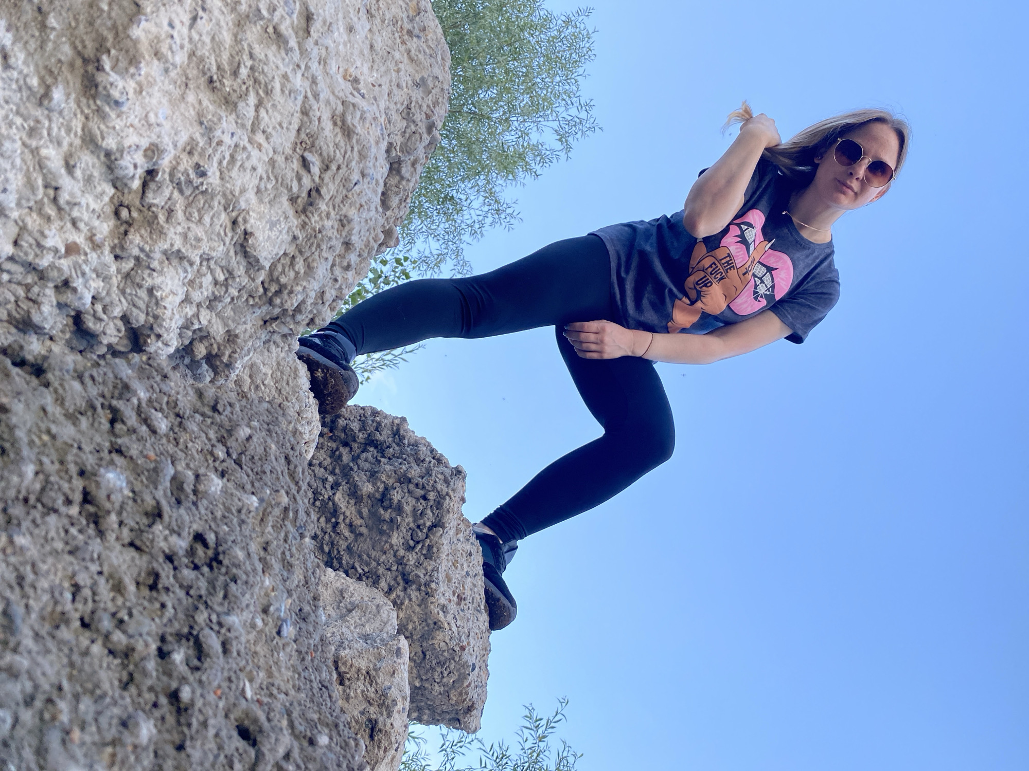 In an outdoor, low-angle shot, a woman stands on large, rough, light-colored concrete structures against a clear blue sky. She is wearing dark leggings, a dark t-shirt with a graphic design of lips and text, and sunglasses. Her left leg is bent and positioned forward, while her right leg is extended back, and she is leaning slightly to her left, touching her hair with her right hand. The concrete structures appear to be weathered and have a coarse texture, with smaller pieces of debris scattered around. The background consists of the sky and some distant greenery. The location is identified as Chaikovsky, Russia.