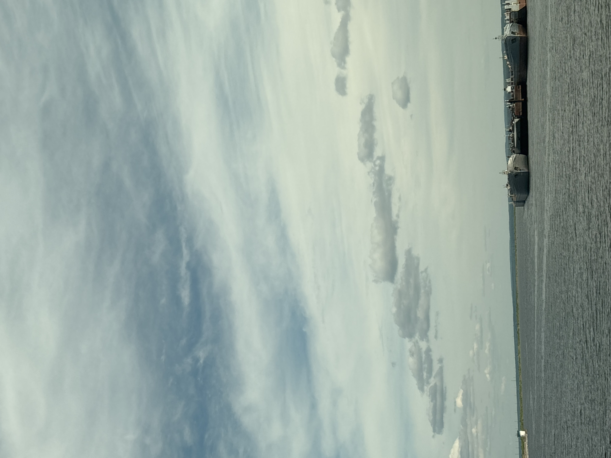The image displays a vast waterscape under an expansive sky, likely a view across a wide river or large lake. The sky occupies the upper two-thirds of the frame, filled with a diverse cloudscape. On the left, wispy, fibrous clouds stretch across the sky, varying in density and creating a diffused, almost ethereal effect. Towards the right, more distinct, puffy cumulus clouds are scattered, casting subtle shadows and adding texture to the sky. The overall color of the sky is a mix of light blue and various shades of grey-white, indicating a partly cloudy or overcast day.
The lower third of the image features the wide body of water, appearing calm with gentle ripples on its surface, reflecting the muted tones of the sky. Along the distant right bank, a series of large, dark-hulled vessels, possibly cargo barges or industrial boats, are lined up, seemingly docked or anchored. Their superstructures are lighter in color, standing out against the darker hulls and the distant shoreline. A faint green strip of land is visible along the horizon line, separating the water from the distant sky.
There are no people visible in the scene, and no specific activities are explicitly depicted; the boats appear stationary. The lighting suggests it is daytime, with soft, diffused light, indicating mild weather conditions. Given the provided location context, this scene likely depicts a section of the Kama River in Chaikovsky, Russia. The image evokes a sense of expansive tranquility with an underlying industrial presence. No text is visible within the image.