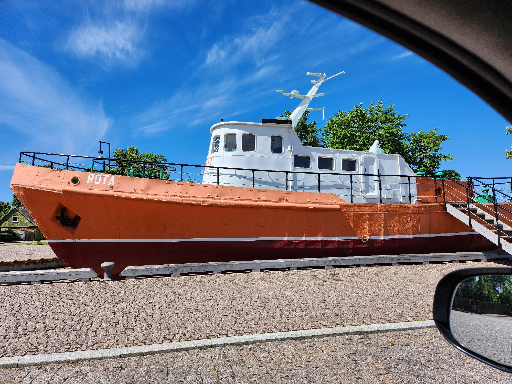 An orange and white ship named ROTA is docked on a paved area under a bright blue sky with scattered white clouds. The ship is positioned horizontally, occupying the majority of the frame. The front of the ship is to the left, featuring an anchor. The ship has a white cabin with several windows and a radar antenna on top. A set of stairs leads up to the deck on the right side of the ship. Trees and a building are visible in the background. The foreground consists of a cobblestone surface, and a car's side mirror is visible in the lower right corner, reflecting a street and trees.
