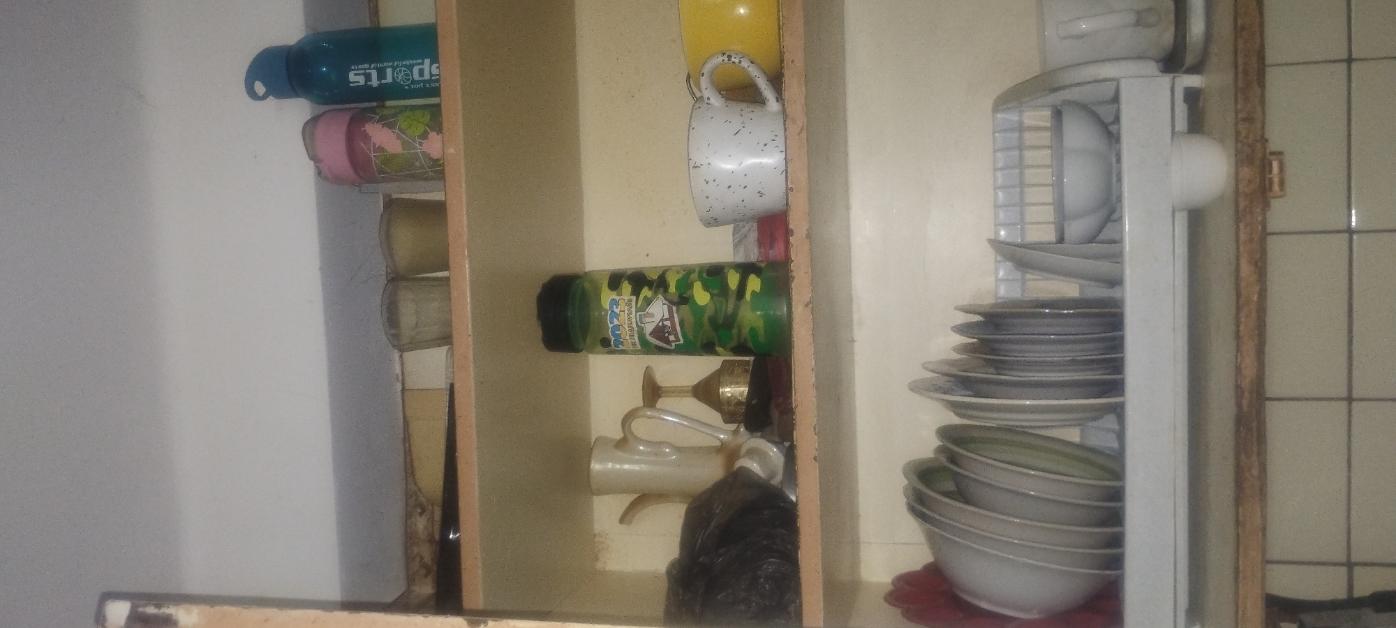 The image shows a close-up, slightly angled view of a kitchen shelf unit in Zaporizhzhia, Ukraine. The unit appears to be made of wood with a faded, beige finish. The shelves are sparsely stocked.
On the top shelf, from left to right, are two clear, ribbed glasses, a pink and green patterned cylindrical container, and a teal blue water bottle with "SPORTS" written on it.
The middle shelf contains a light-colored jug with a handle and spout, a gold-colored trophy-like object, and a green camouflage-patterned water bottle with a black lid, featuring a house and "2023" printed on it. To the right of the water bottle are a speckled white mug with a handle and a yellow mug or bowl. A black plastic bag is positioned to the left of the jug.
The bottom shelf holds a white dish rack containing several stacks of plates and bowls. To the right of the dish rack, a white cup with a handle is visible, and below the dish rack, a small white bowl sits on the shelf.
The background appears to be a tiled wall. The overall impression is of a utilitarian kitchen space.