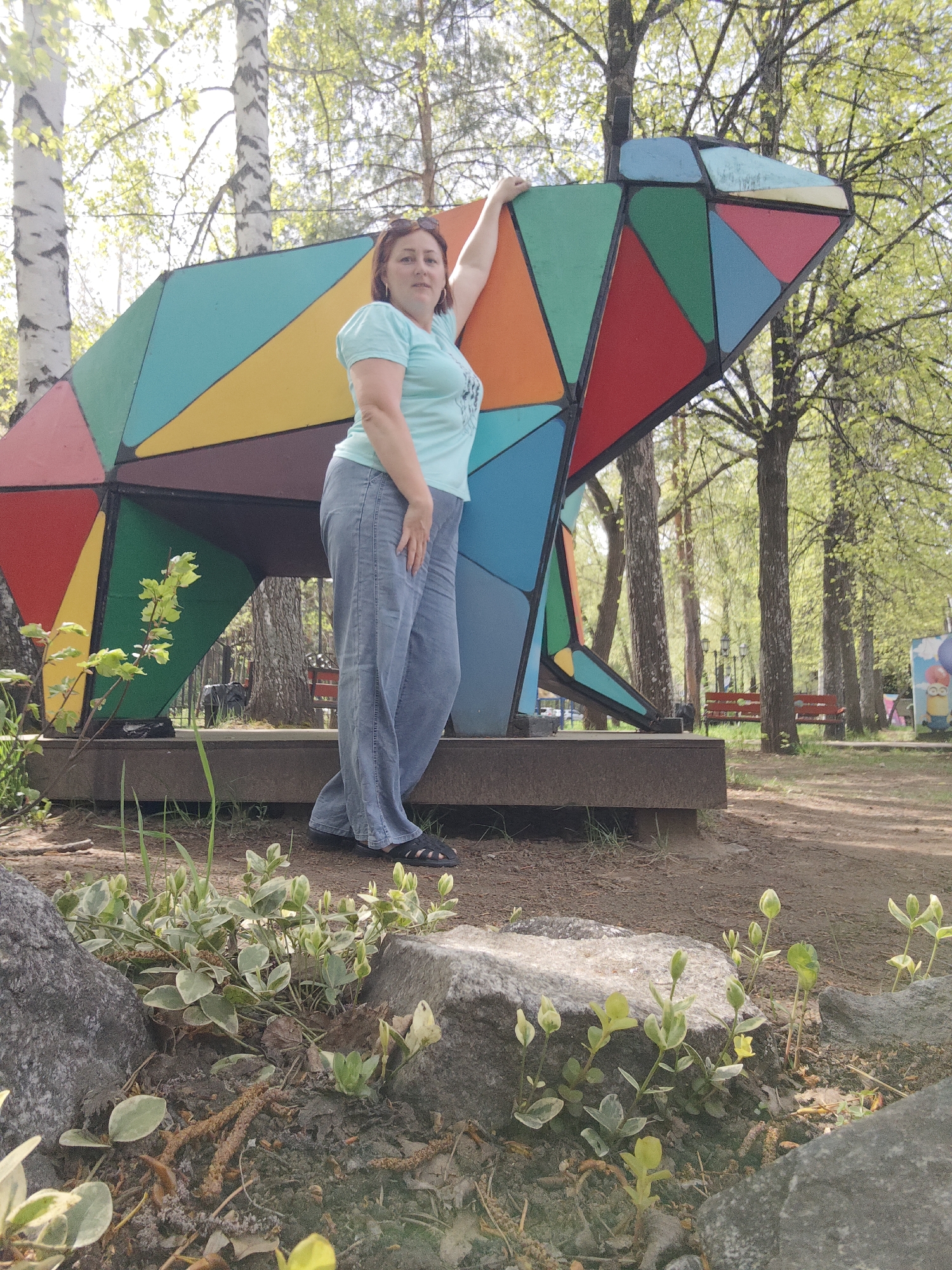A woman stands next to a large, polygonal, multi-colored bear sculpture in a park-like setting. The sculpture is elevated on a concrete base. The woman has shoulder-length red hair and wears a light blue t-shirt and blue jeans. She leans on the bear sculpture with her left arm. Behind the woman and the sculpture are trees with light green leaves and several birch trees. In the background, benches can be seen. The foreground features a small rock with some leafy vegetation. The location is the city of Chaikovsky, Russia.