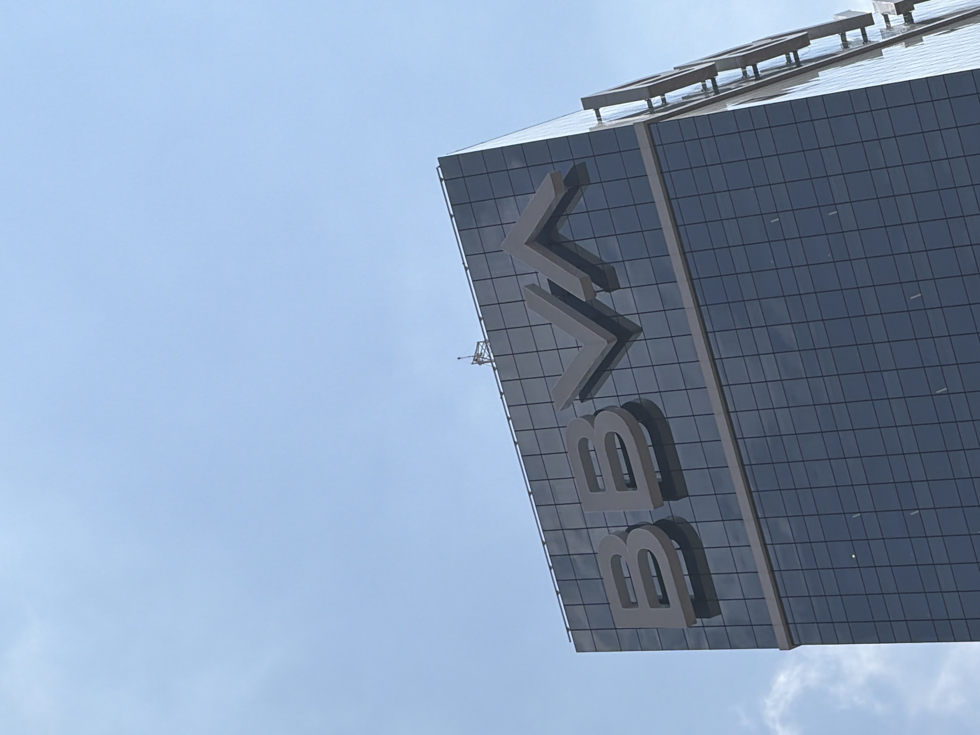 The image shows a low-angle shot of a building with the "BBVA" logo on its side, likely the BBVA Tower in San Bernardino Tlaxcalancingo, Mexico. The building is modern, clad in dark glass panels forming a grid-like pattern. The logo is large and grey. A communication tower stands on top of the building. The sky is light blue.