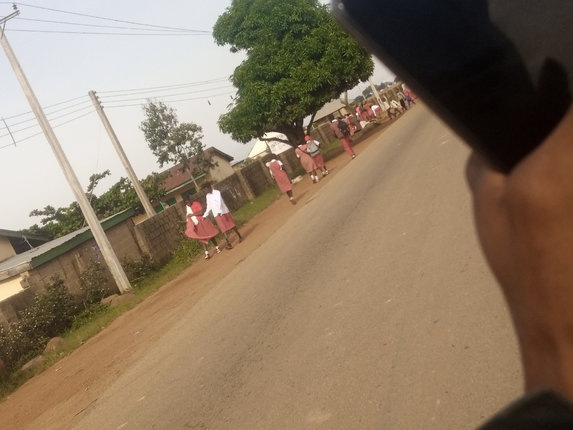 The image shows a street scene in Katon Rikkos, Nigeria. Several people, mostly young individuals in school uniforms, are walking along the side of a paved road. The uniforms appear to consist of white shirts and red skirts or shorts. Some of them are carrying backpacks, suggesting they are likely students.
The setting includes a brick wall bordering a property, with some greenery and trees visible behind it. Two prominent utility poles with overhead wires stand on the left side of the road, indicating the presence of electrical infrastructure. A large, leafy tree dominates the background on the right, providing shade. The sky is overcast, suggesting a diffused light, possibly late afternoon or early morning.
The overall atmosphere seems casual, with people moving along the road, some in small groups. The activity appears to be a daily commute or a gathering of students. There is no visible text in the image. The perspective suggests the photo was taken from a moving vehicle or by someone holding a phone close to the camera.
