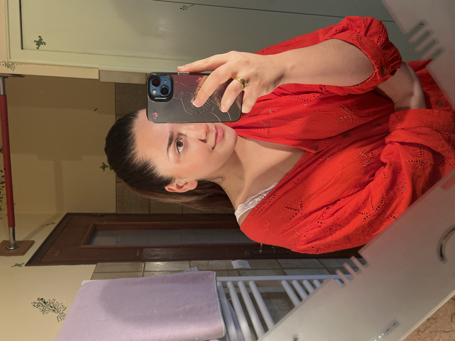A female individual, aged approximately 20-30 years, is depicted taking a mirror selfie in a bathroom located in Ancenis, France. Her dark hair is pulled back into a ponytail. She is wearing a red, eyelet-embroidered, long-sleeved shirt, partially unbuttoned to reveal a white lace camisole or bra strap. She holds a dark-colored smartphone in her right hand, which obscures the right side of her face; her left eye and the left side of her face are visible, showing a slight smile. A gold ring with a small clear stone is present on her left ring finger. The background, reflected in the mirror, consists of light yellow walls adorned with green stenciled botanical and frog motifs. A dark brown wooden door is partially visible on the left. Above the door, a red cylindrical bar is mounted. A white, multi-barred towel warmer, over which a light purple towel is draped, is visible on the far left of the reflection. The mirror itself has an etched design along its bottom edge.
