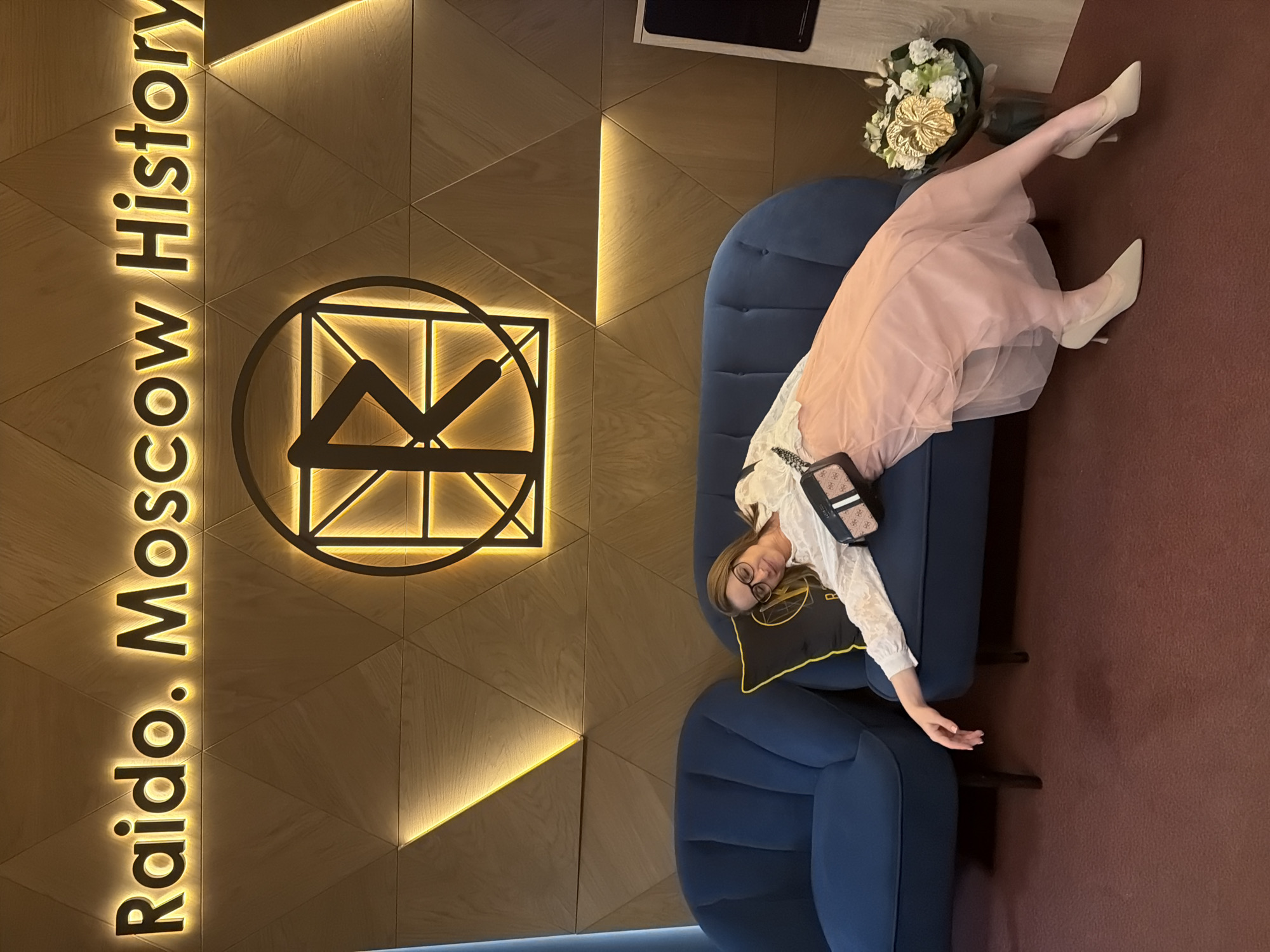 A young woman lies prone on a blue velvet sofa in a modern, illuminated interior. She wears a white, lace-trimmed shirt, a pale pink skirt, and light-colored high heels. A small, patterned purse lies near her, and a pillow is situated beneath her head. The background includes a geometrically patterned wall with an illuminated logo and the words "Raido. Moscow History." This suggests the location is likely a Raido restaurant or establishment located in Moscow, although Khimki is not visible. A bouquet of flowers sits nearby on a table, and a portion of a television is visible in the upper background. The floor is brown, and the overall setting appears stylish and contemporary.