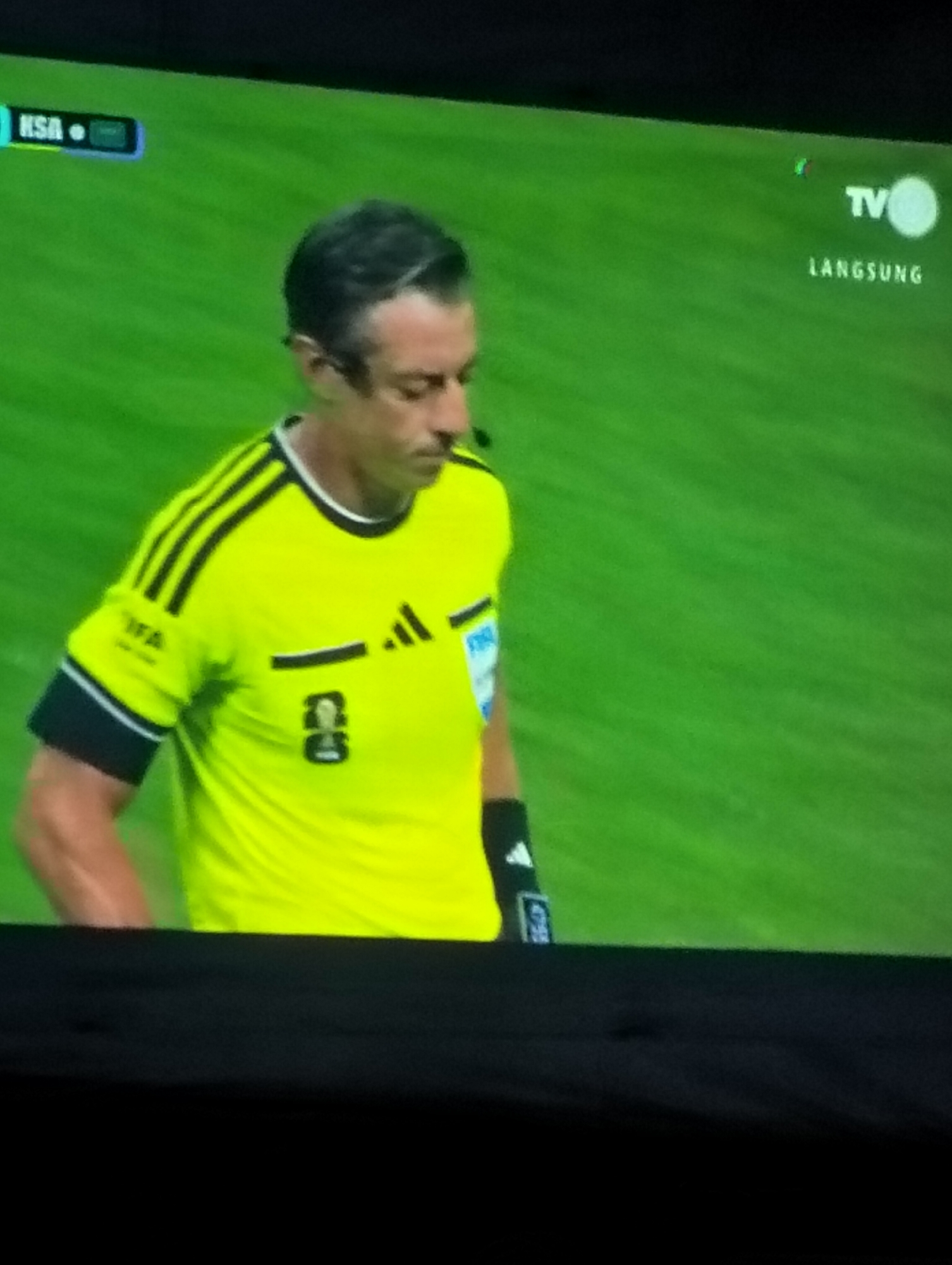 The image shows a soccer referee on a bright green field. The referee is wearing a neon yellow jersey with black stripes on the sleeves and black trim on the collar. The jersey has the "FIFA" logo on the left sleeve and an Adidas logo on the right chest. There is a number "8" printed on the front of the jersey. The referee has dark hair and is looking down. On the left side of the screen, there is a blue scoreboard-like graphic with the text "KSA". On the right side of the screen, there is a "TV" logo with a circular graphic and the word "LANGSUNG" written below it. The background is a green, out-of-focus soccer field. The image is a digital capture, likely from a television broadcast, with the bottom portion of the screen obscured by a black border. The location in Payagaboh, Indonesia is not discernible from the provided image.
