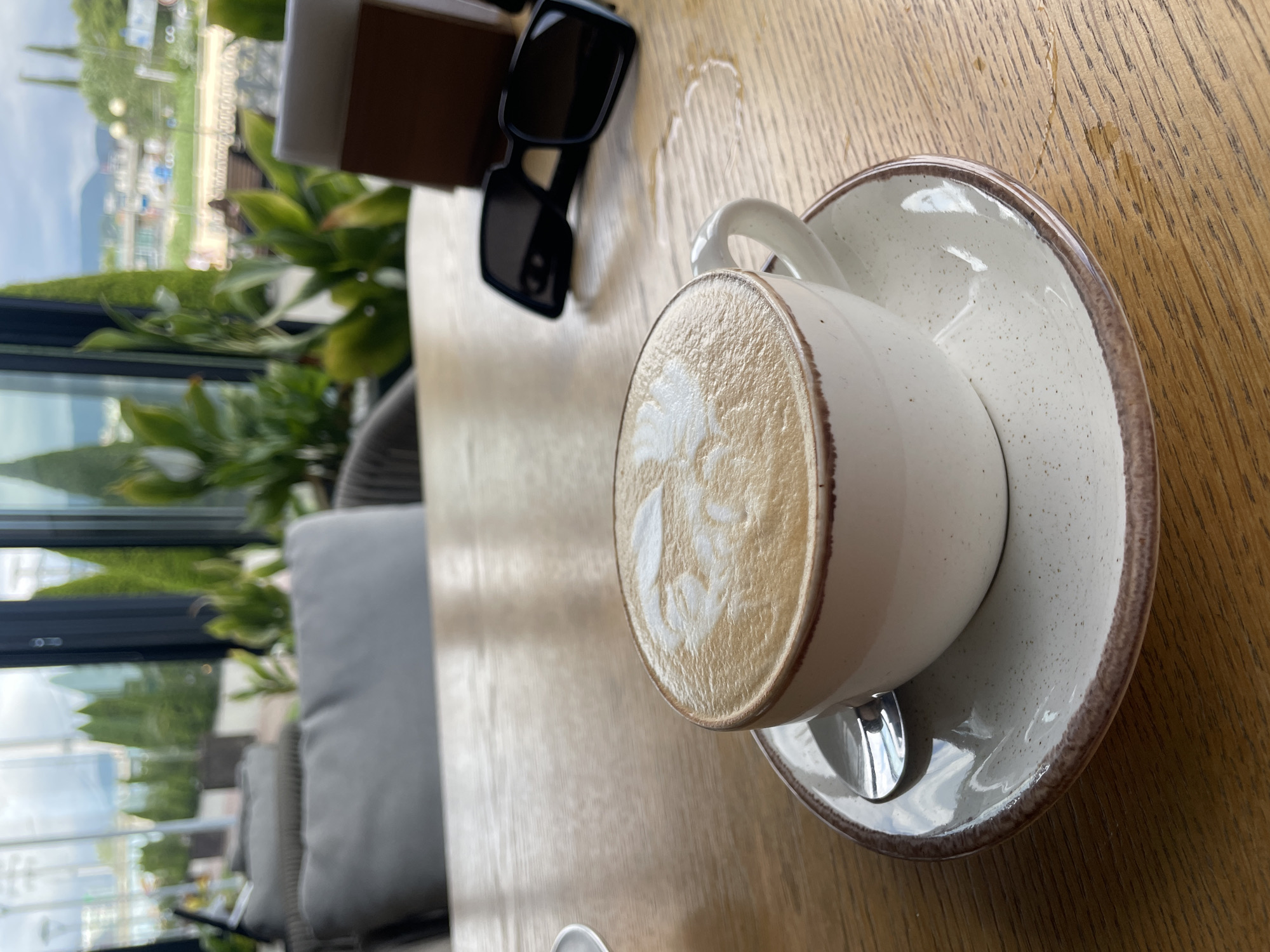A close-up, overhead shot shows a white ceramic cup of latte on a matching speckled saucer, placed on a wooden table. The latte features intricate milk foam art. To the left of the cup, a pair of black sunglasses rests on the table, partially obscuring a brown paper container. In the background, a blurred view through a window reveals lush green foliage and a glimpse of buildings and distant mountains, suggesting an outdoor seating area with a scenic backdrop. The lighting indicates it is daytime, possibly late morning or afternoon. The scene appears relaxed and casual, possibly a moment of leisure during a meal or break. There is no visible text in the image. The overall impression is one of enjoying a beverage in a pleasant, possibly resort-like, setting.