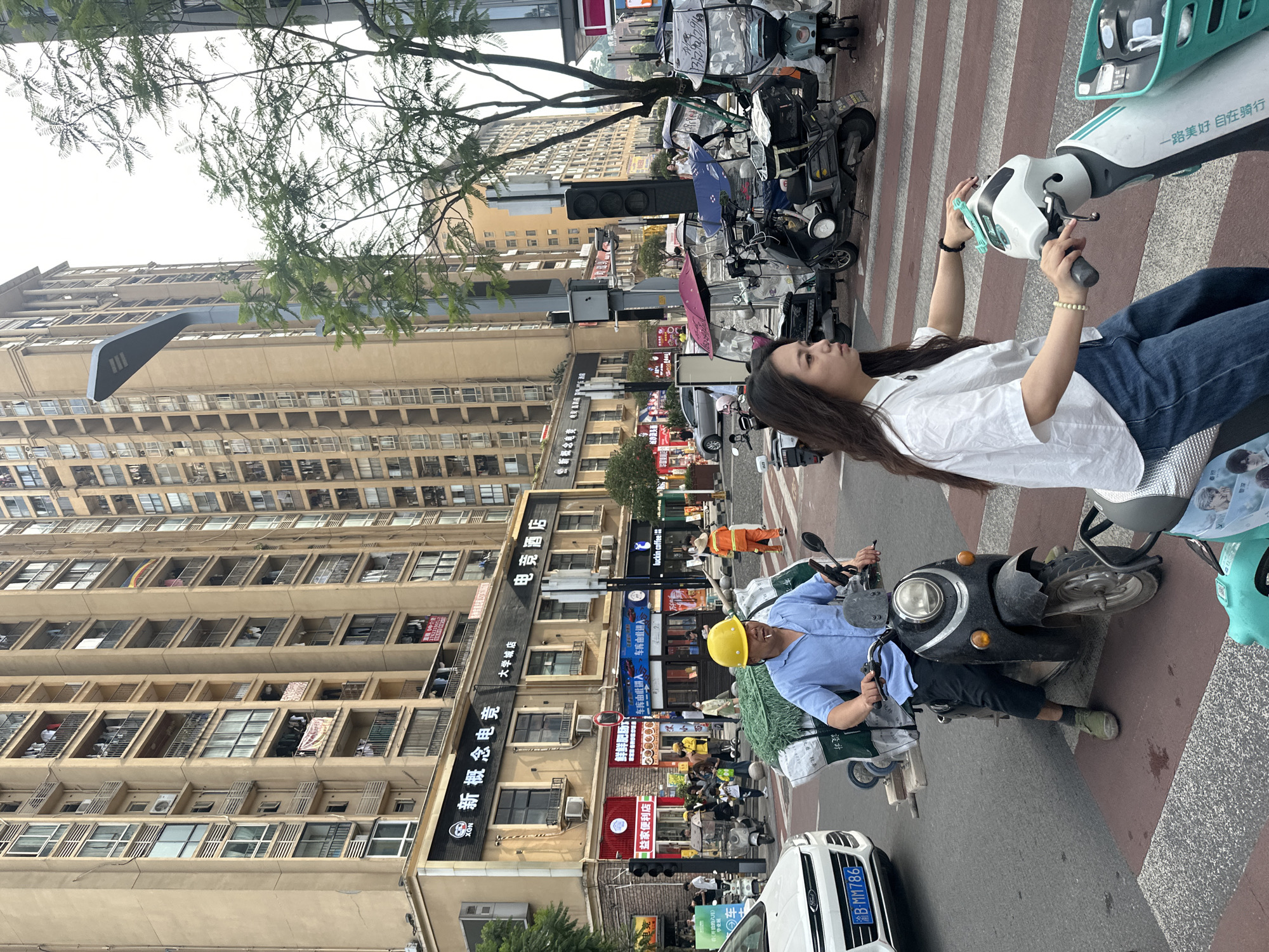 In Chongqing High-tech Zone, China, a man wearing a blue shirt and a yellow hard hat rides a dark-colored scooter loaded with green netting and what appear to be bundled goods. He is in a crosswalk, with a white car to his left and other scooters to his right. A woman in a white shirt and blue jeans is on a light-colored scooter, also in the crosswalk, looking to her left. Behind them, tall residential buildings line the street, with shops and businesses at the ground level. A street light with multiple lamps stands near the intersection. The scene depicts urban traffic and pedestrian activity.