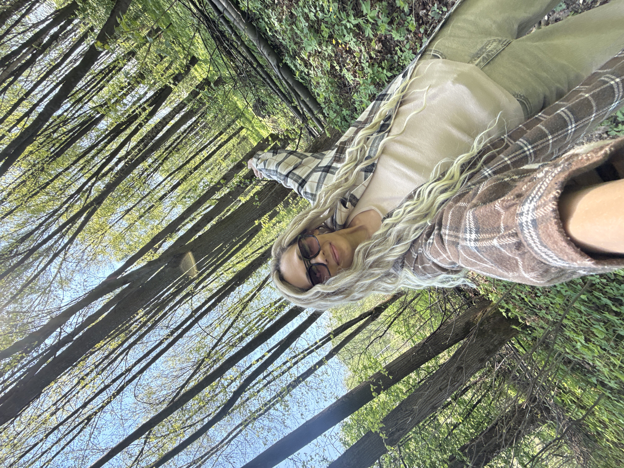 A woman with blonde hair is in a forest setting. She is wearing sunglasses, a light-colored top, a plaid shirt, and jeans. She appears to be taking a selfie. The background consists of tall trees with green foliage and a glimpse of the sky. The exact location within the city of Rusanovo, Russia, cannot be determined from the image.