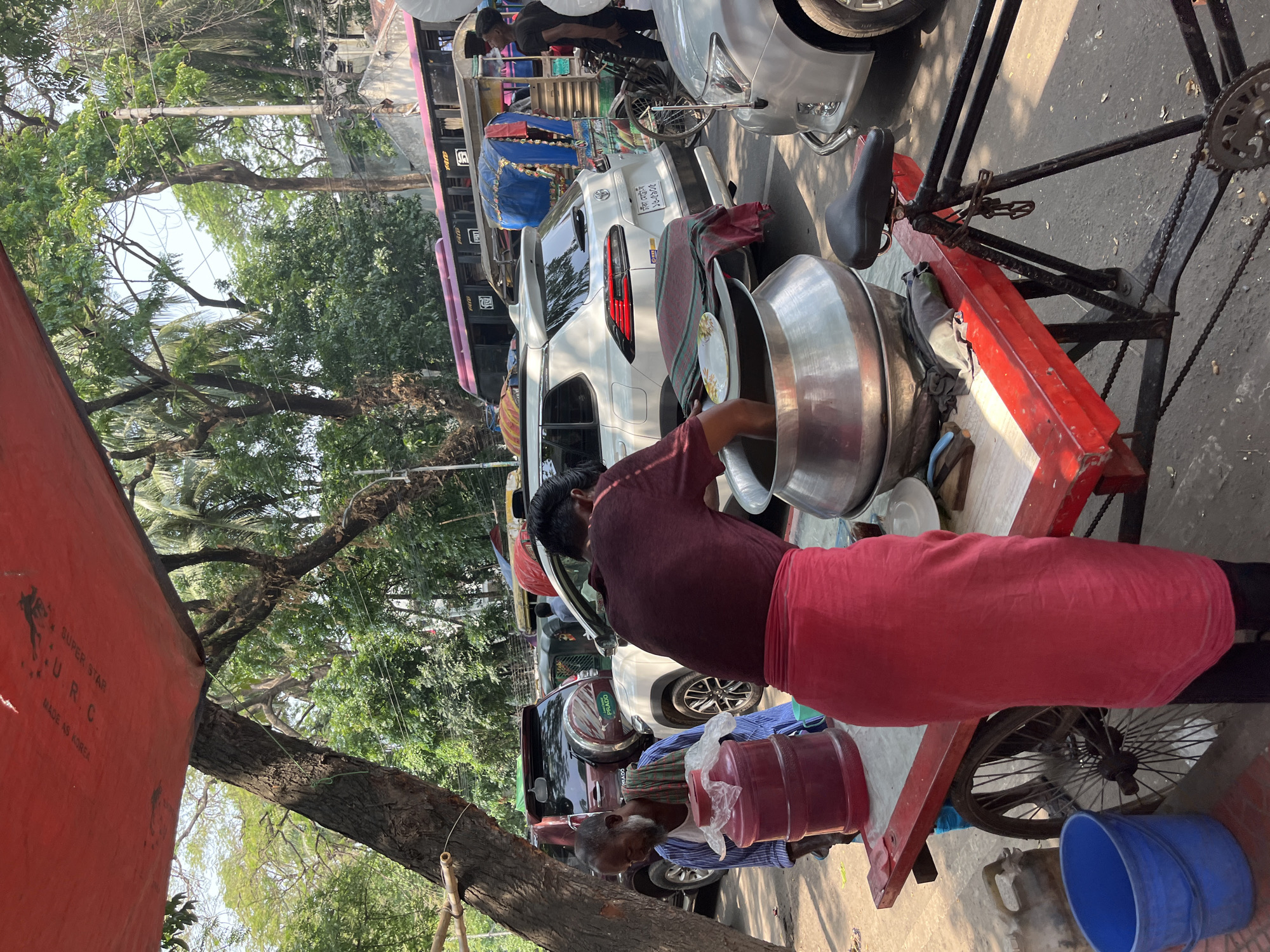 A man wearing a maroon shirt and red lungi is serving food from a large aluminum container on a red cart attached to a bicycle in Dhaka, Bangladesh. An elderly man with a gray beard sits nearby, holding plastic containers. The scene is a busy street with trees, cars, buses, and other vehicles visible in the background. The word "URC" and "MADE AS KOREA" are partially visible on a red structure.