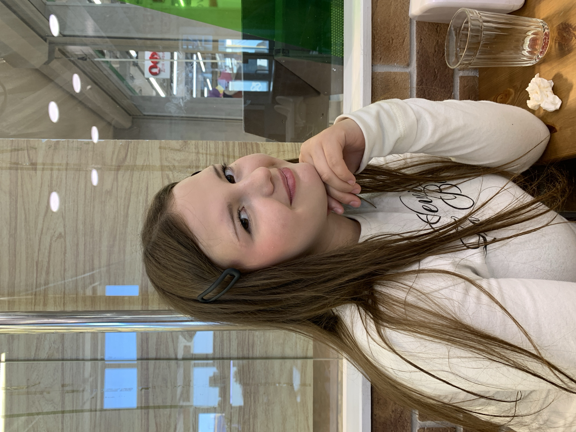 A young woman with long brown hair and a black hair clip is seated at a wooden table, resting her chin on her hand and looking towards the viewer with a slight smile. She is wearing a white long-sleeved shirt with a graphic design. In the background, through a glass partition with reflective surfaces, a retail environment with shelves and signage is visible. A clear glass, a crumpled white napkin, and a brick-patterned wall are on the table in front of her. The setting appears to be a cafe or dining area within a public space in Chaikovsky, Russia.