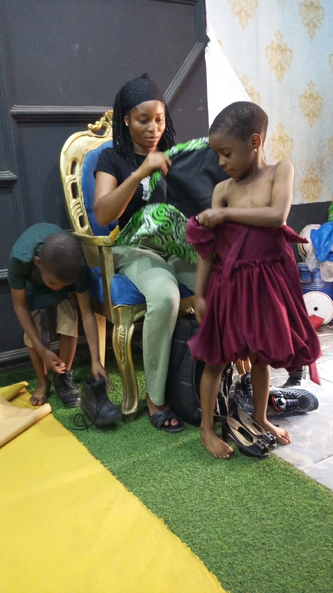 The image shows three people in what appears to be an indoor setting. A woman with braided hair and a black head wrap is sitting on a gold-trimmed chair with a blue seat and back. She is wearing a black shirt and light green pants, and she is holding up what seems to be a black and green patterned piece of fabric. To the left of the chair, a young boy in a green shirt and khaki shorts is bending over, appearing to put on a black shoe or boot. To the right of the chair, a young, dark-skinned child is standing partially undressed, holding a maroon or dark red dress. Shoes and boots are scattered on the floor around the child, near the chair.
The floor is divided into two sections: one with artificial green turf and another with a yellow material. In the background, the wall has a patterned wallpaper design, and there is a dark panel or piece of furniture adjacent to the chair.
The location is identified as Unknown, Unknown.