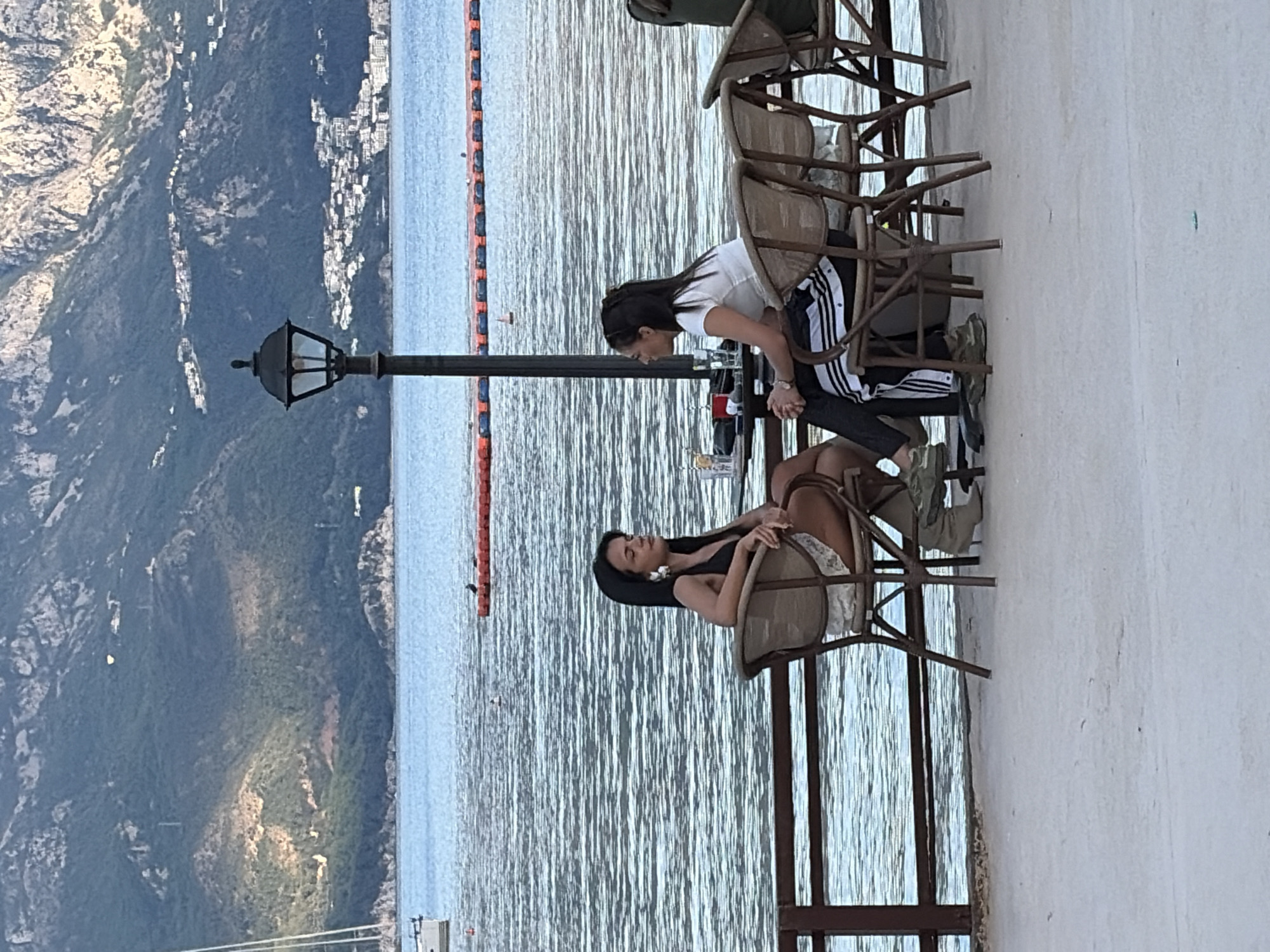 Two women are interacting at an outdoor cafe located in Budva, Montenegro. The cafe is situated alongside a body of water, with mountains visible in the background. One woman is sitting on a cafe chair while the other is leaning forward to converse with her. Several empty cafe chairs are nearby. A lamppost stands close to the women.