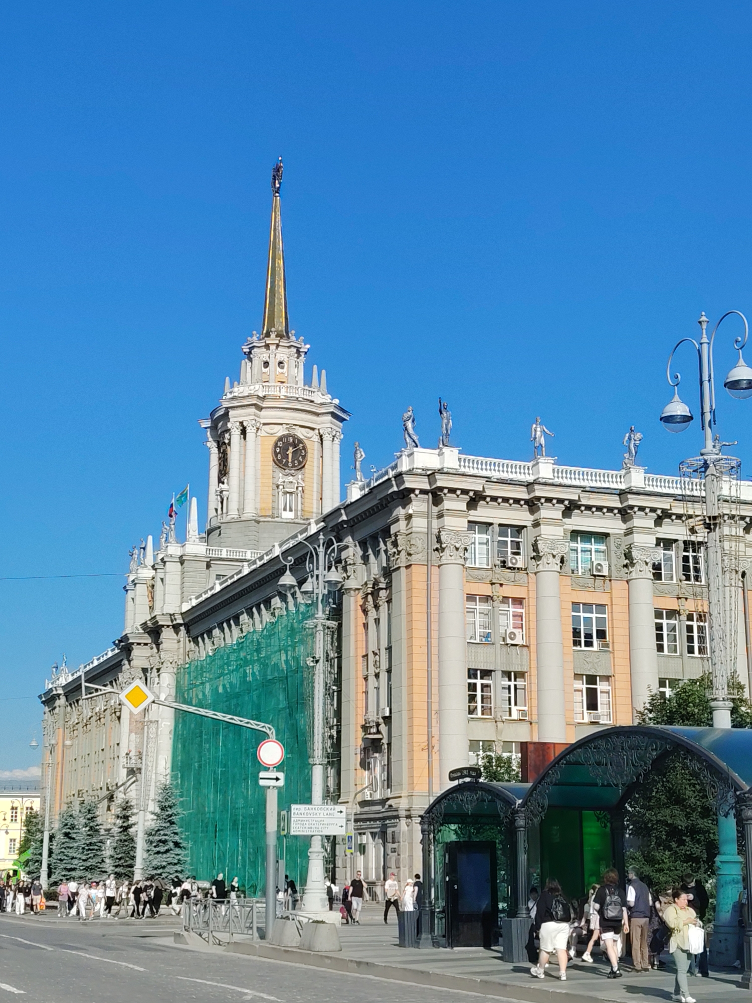 The image captures a sunny day in Yekaterinburg, Russia, featuring a grand, ornate building with a prominent clock tower. The architecture is classical, with columns, statues adorning the roofline, and decorative details. The building is partially covered in green scaffolding, suggesting ongoing renovation or construction.
In the foreground, a street scene unfolds with a clear blue sky overhead, indicating good weather. A group of people are walking on the sidewalk and crossing the street, casually dressed and appearing to be going about their day. To the right, an elaborate, arched green structure, possibly a bus stop or public art installation, is visible.
A street sign in the image reads "ОРД. БАНКОВСКИЙ ЛАЙН" and "BANKOVSKY LANE," providing a specific location clue. The time on the clock tower appears to be around 10:10. The overall atmosphere is one of an active urban environment on a pleasant day, with a blend of historical architecture and modern life.