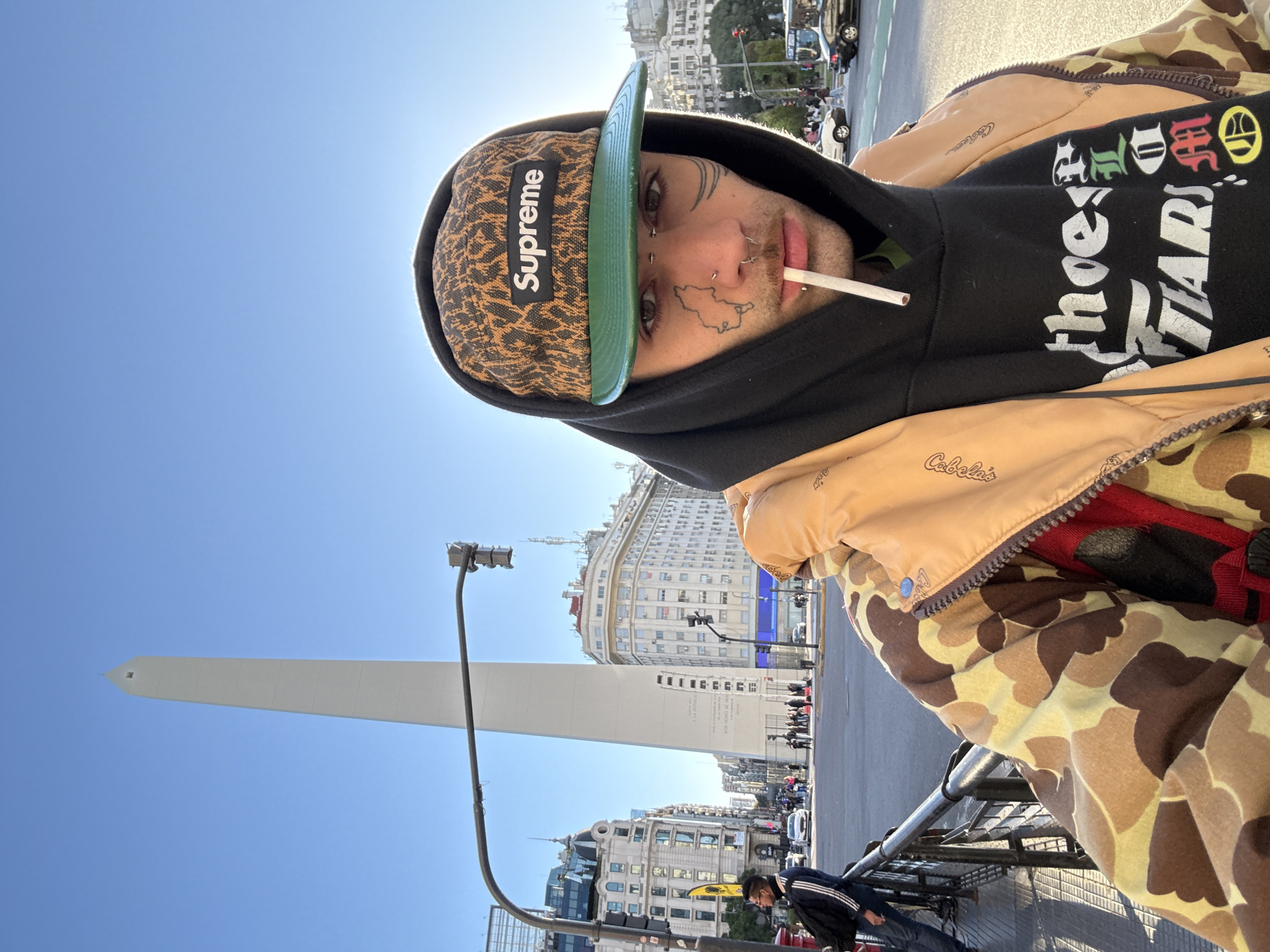 The photograph features a person taking a selfie in Buenos Aires, Argentina. The person is wearing a Supreme hat, a black hoodie, and a camouflage jacket. They have face tattoos and a cigarette in their mouth. Behind the person is the Obelisco de Buenos Aires, a historical monument. The scene is set during the day with clear skies. Other buildings, traffic, and pedestrians are visible in the background.