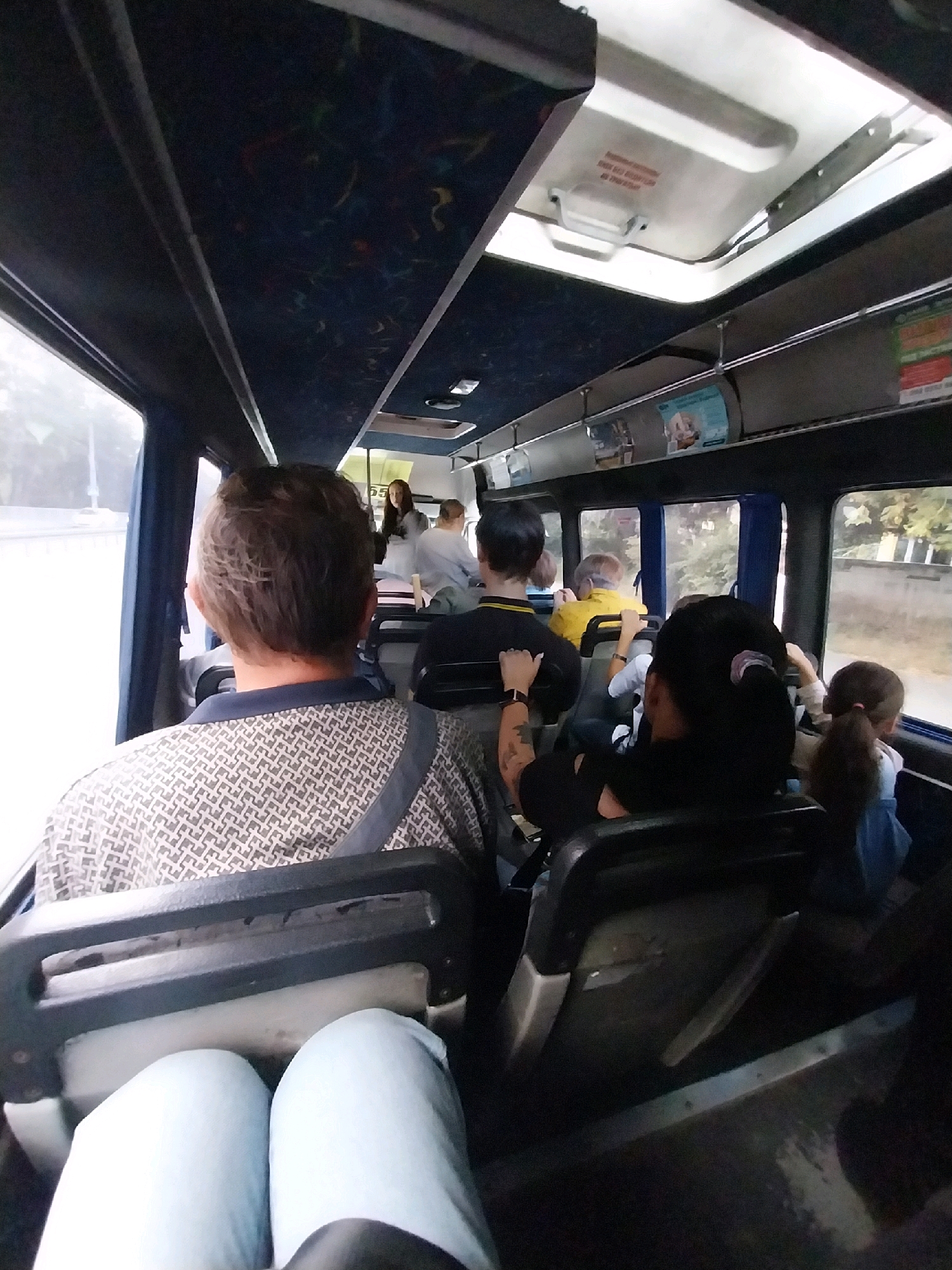 The image depicts the interior of a public bus with several passengers seated. The perspective is from a passenger looking forward and slightly to the right. The bus has blue-tinted windows, dark blue upholstery on the ceiling with a pattern, and visible overhead storage racks. Light filters in from the windows, indicating it is daytime.
In the foreground, a person is seated, facing forward, wearing light blue pants. The back of their head and shoulders are visible. The seat in front of them is dark gray with a metal safety bar.
Further ahead, other passengers are visible. On the left, a man with brown hair is seated, looking forward, wearing a patterned shirt. To his right, a woman with dark hair tied back in a ponytail is seated, also facing forward. A child with dark hair in a ponytail is seated to her right, wearing a light blue shirt. In the middle of the bus, a person in a yellow shirt is visible, and further back, a woman in a grey jacket stands near what appears to be the bus driver's area, though the driver is not clearly visible.
The environment outside the bus appears to be a roadside with trees and buildings visible through the windows, suggesting an urban or suburban setting. There are advertisements and signage visible on the interior walls of the bus. The overall impression is a mundane commute on public transportation. The prompt requests to include the city of Dnipro, Ukraine, but there are no visual cues in the image that can definitively place it in Dnipro, Ukraine.
